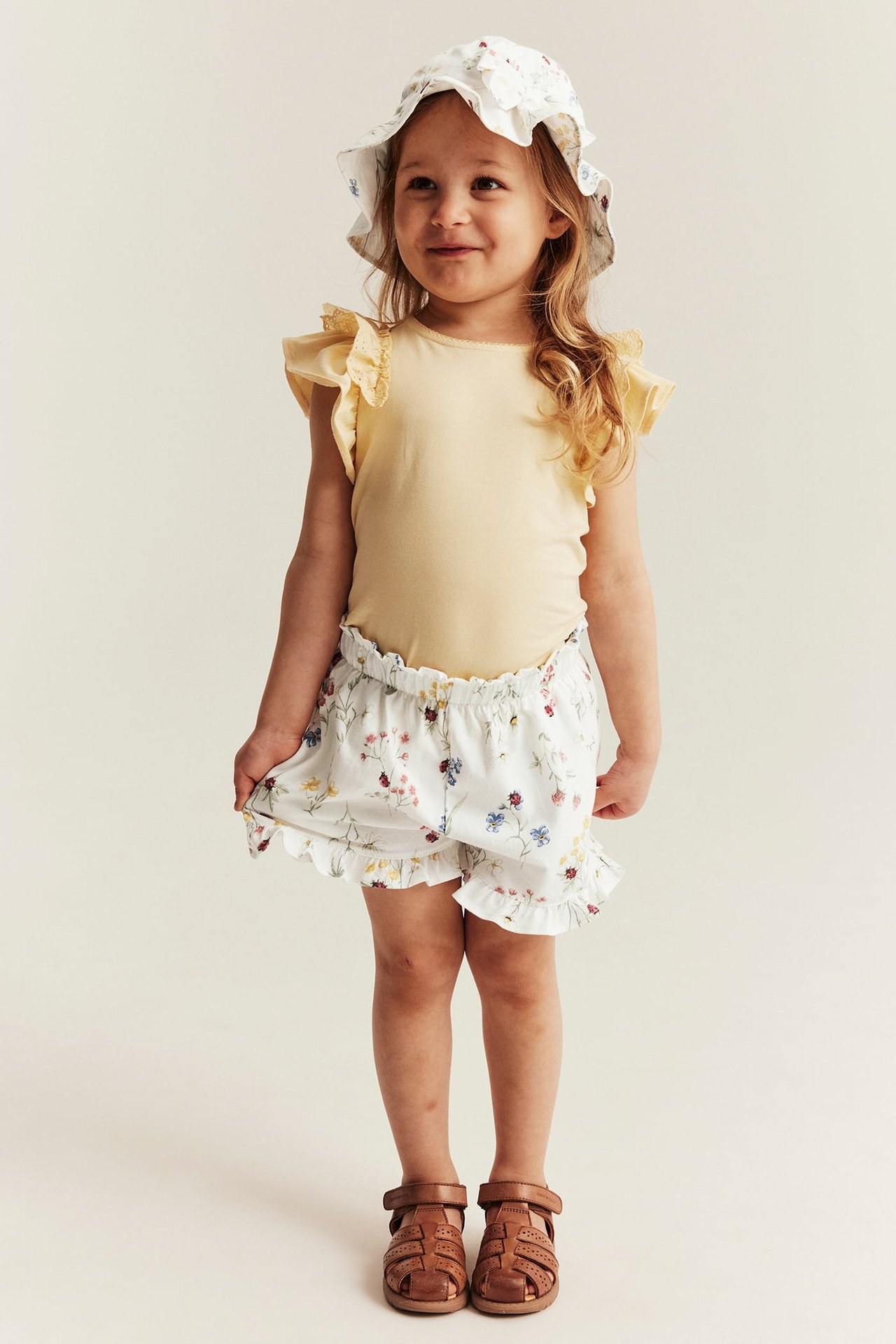 Vorderansicht: Kind trägt offwhite geblümte Jersey-Shorts mit Rüschen, gelbes Oberteil, Blumenhut und braune Sandalen.