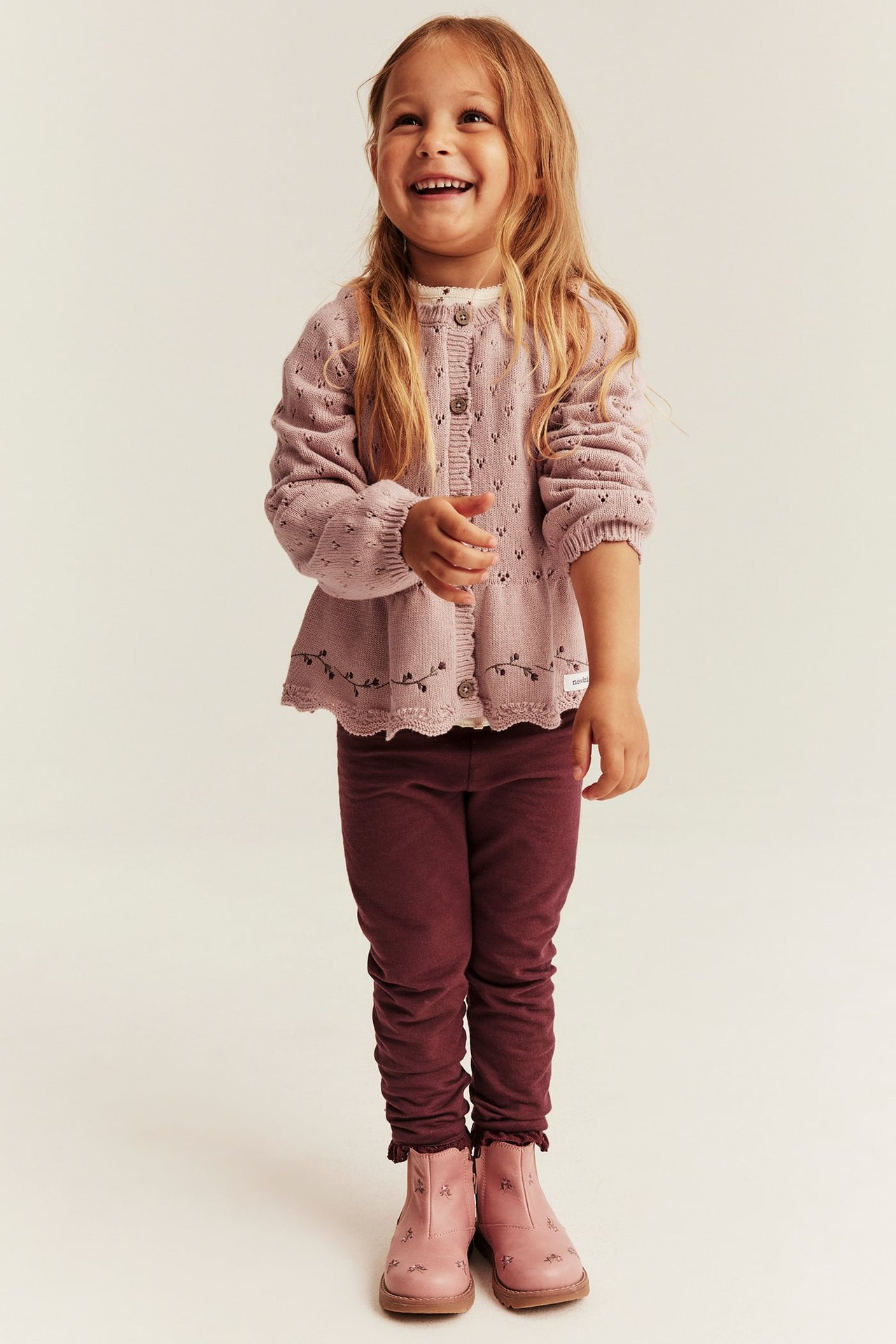 Vorderansicht: Kind in rosa Strick-Peplum-Cardigan, weißem Top, weinroten Leggings und rosa Stiefeln.