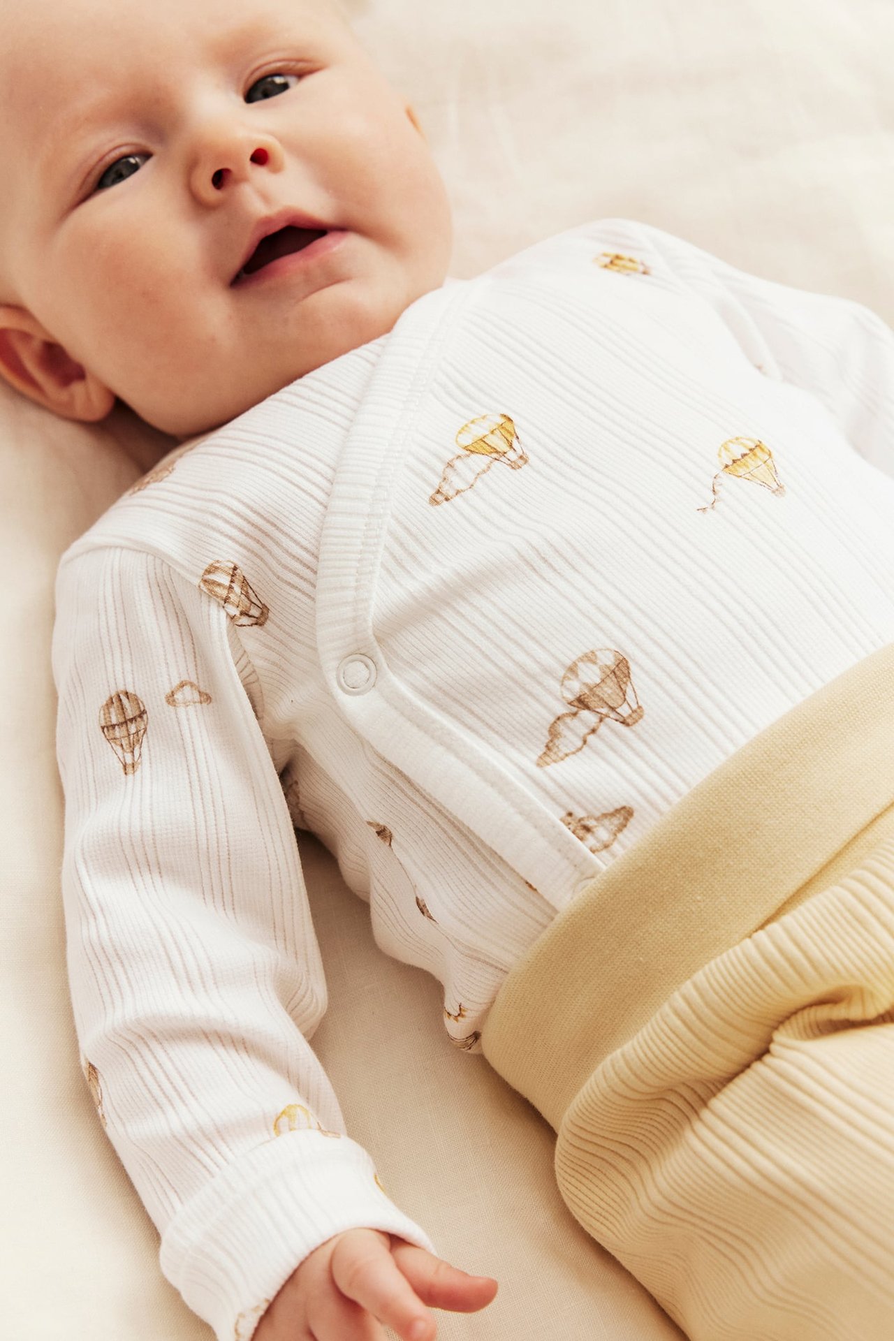 Nahaufnahme Baby in off-white geripptem Wickelbody mit Heißluftballons und gelber gerippter Hose.