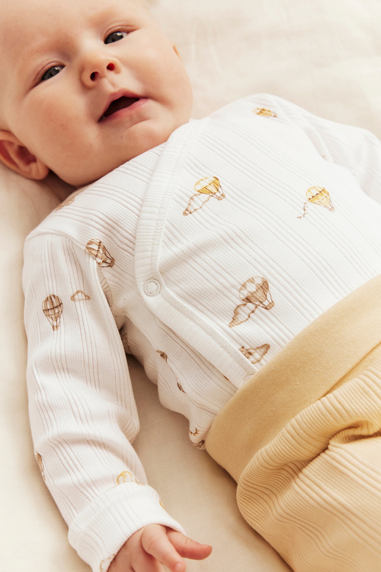 Nahaufnahme von Baby in weißem geripptem Wickelbody mit Heißluftballon-Print und gelber Rippenhose.