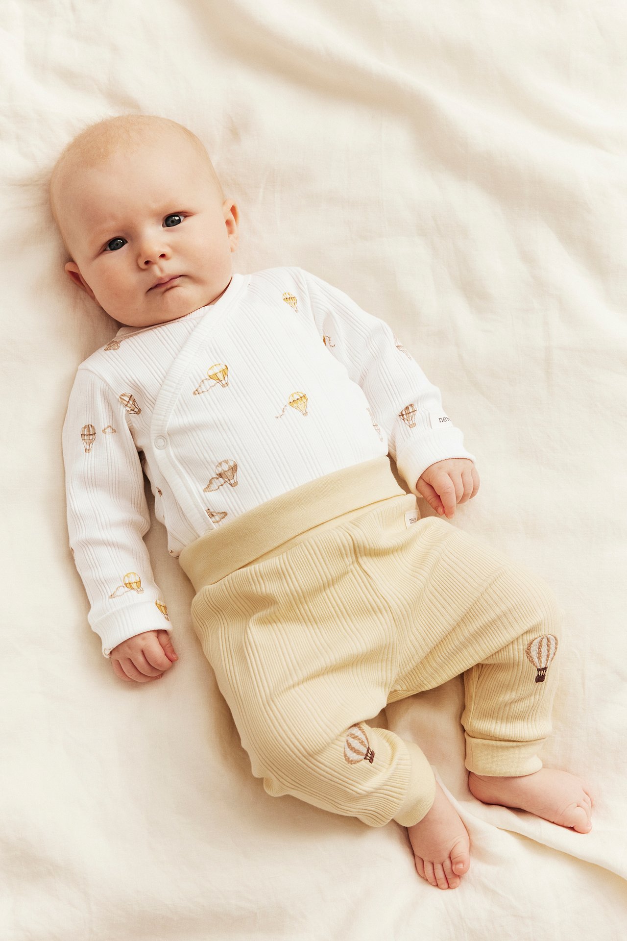 Baby trägt einen cremeweißen gerippten Wickelbody mit Ballonmuster und hellgelbe gerippte Hosen.