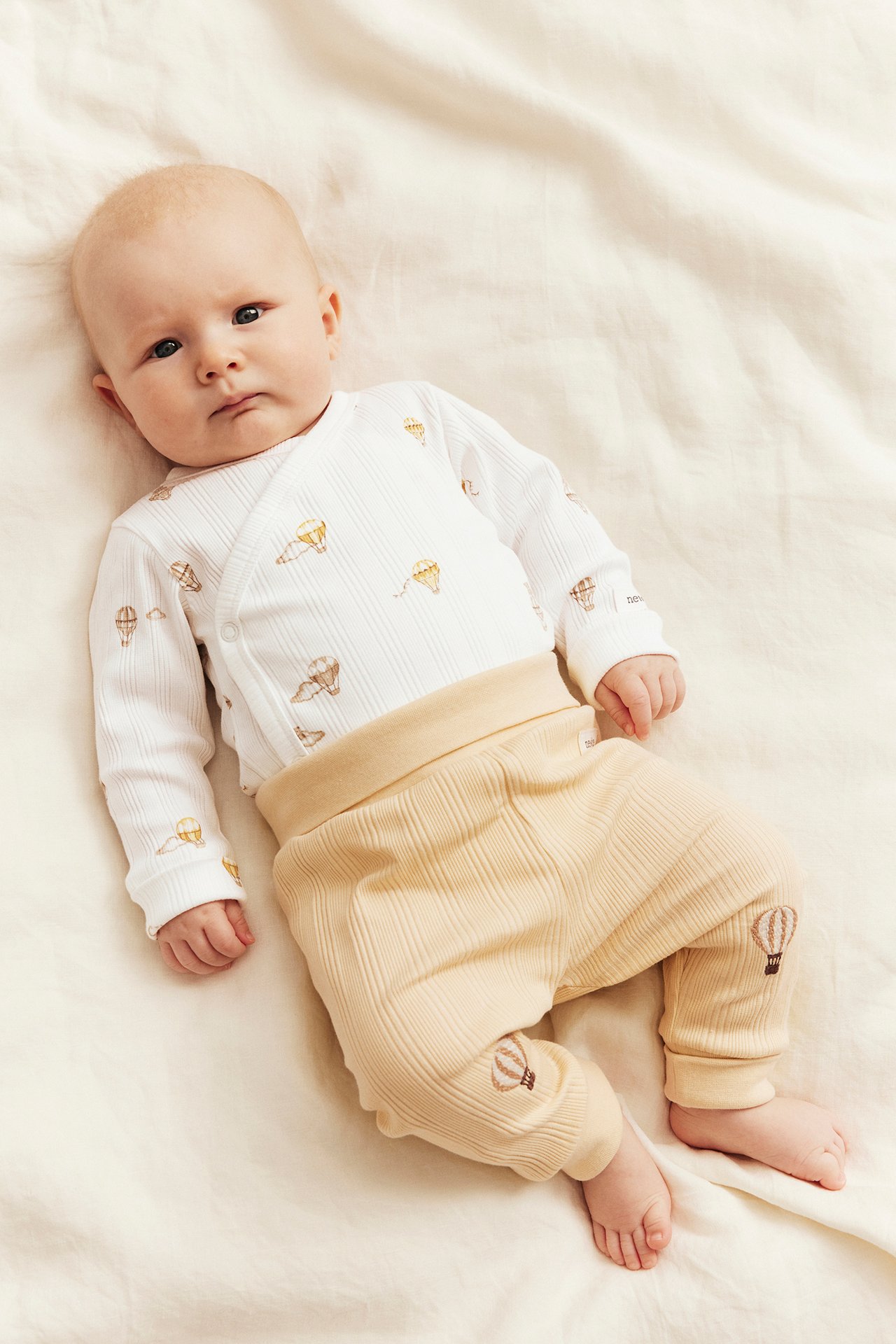 Baby trägt einen weißen gerippten Langarm-Body und eine beige gerippte Hose, beide mit Heißluftballon-Details.