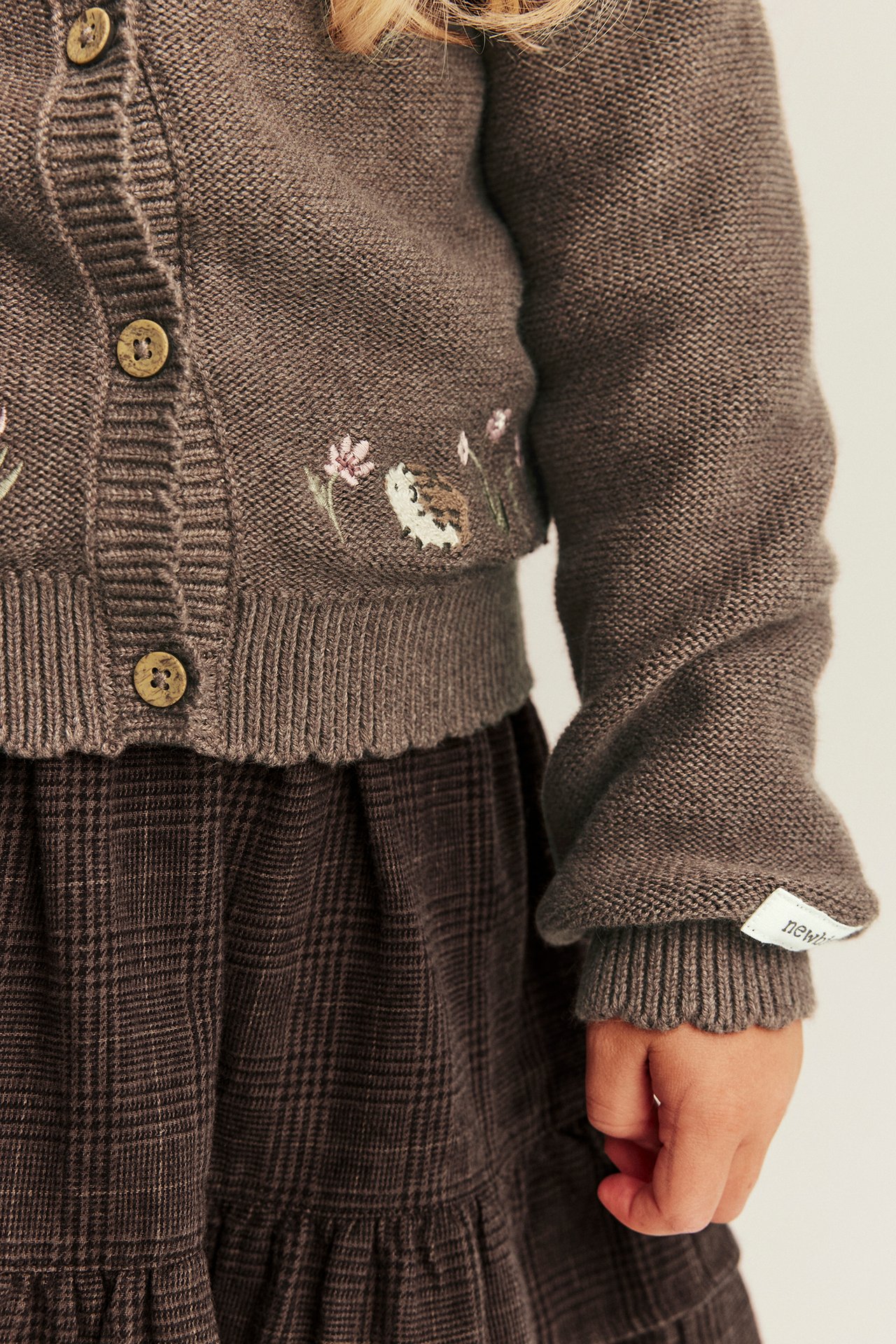 Nahaufnahme: braune Strickjacke mit Igel-Stickerei und braunem Karorock an Kind.