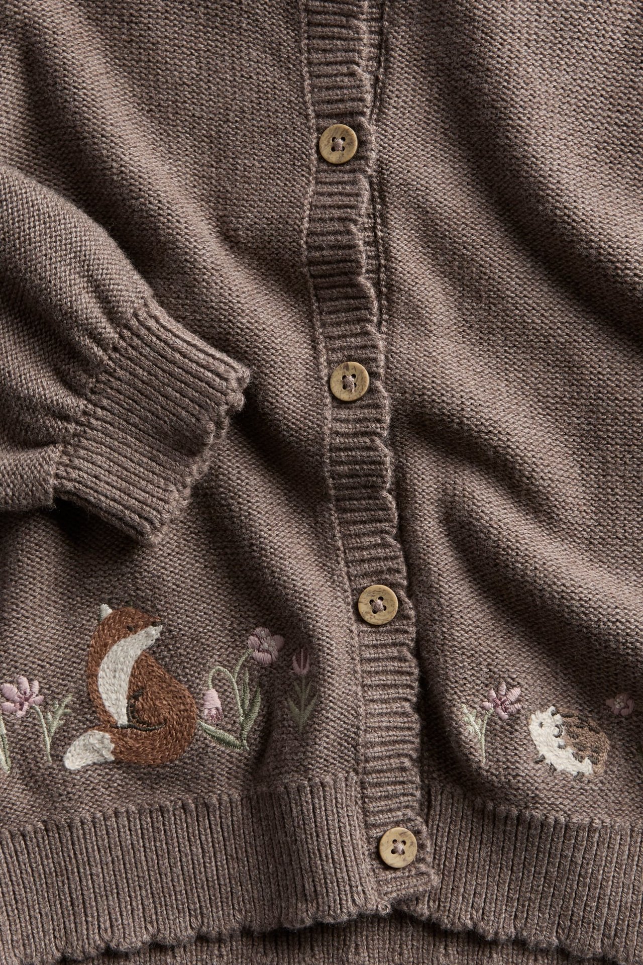Close-up of a brown knitted kid's cardigan with embroidered fox, hedgehog, and wooden buttons.
