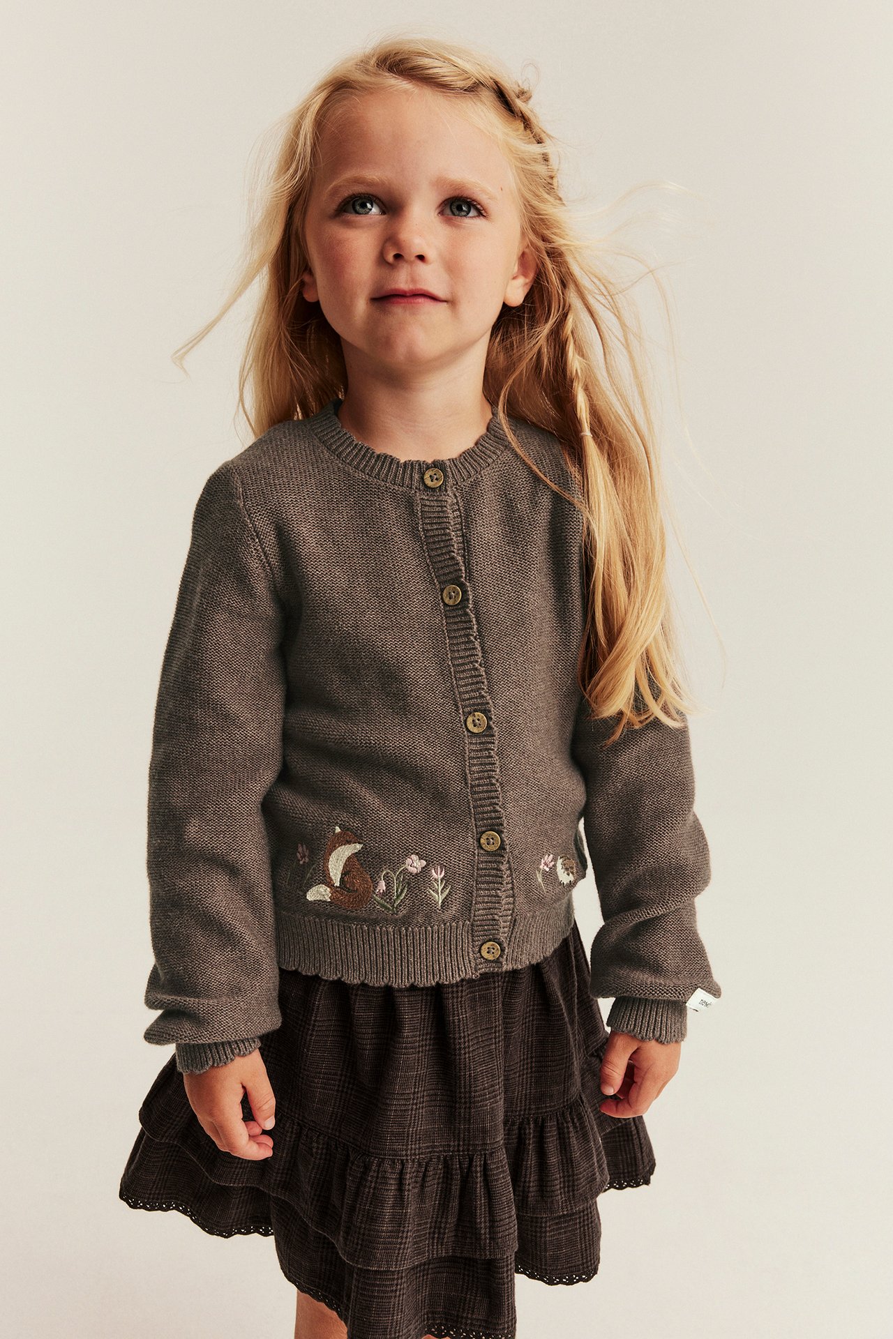 Vorderansicht: Kind trägt braune Strickjacke mit Fuchsbordüre und dunkelbraunen Stufenrock.