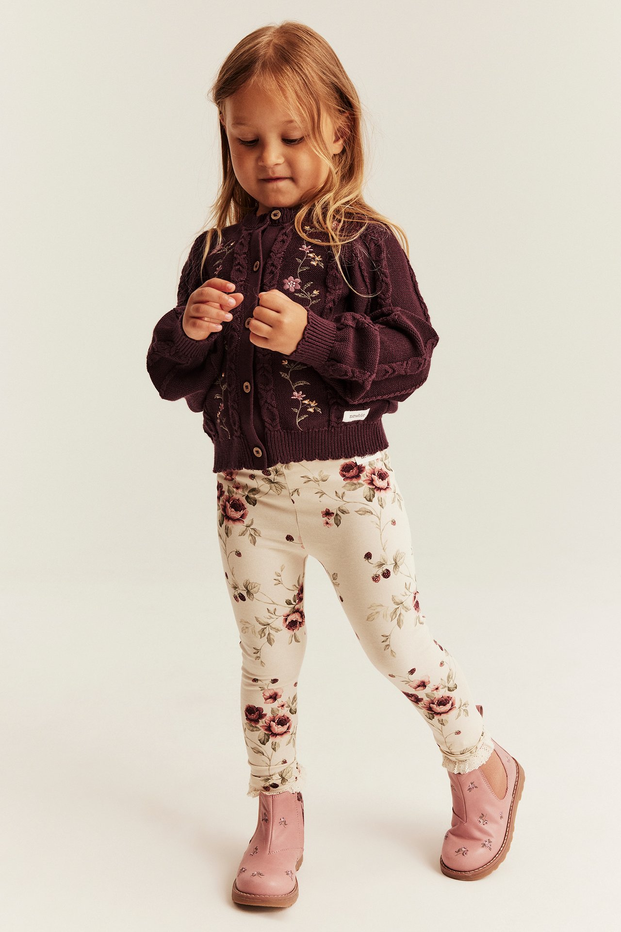 Vorderansicht eines Kindes in dunkelvioletter bestickter Strickjacke, floralen Leggings und rosa Stiefeln.