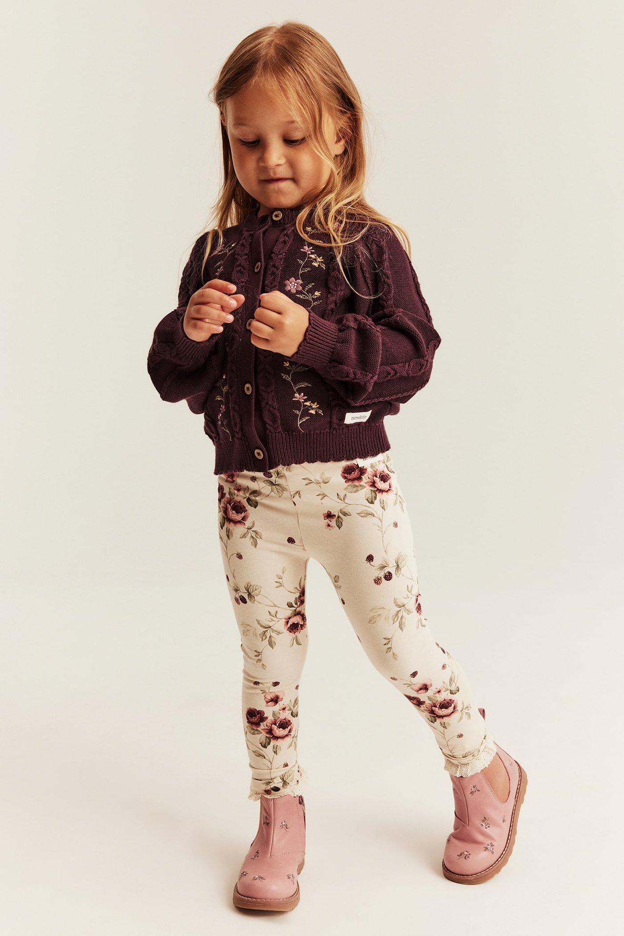 Vorderansicht eines Kindes in dunkelvioletter bestickter Strickjacke, floralen Leggings und rosa Stiefeln.