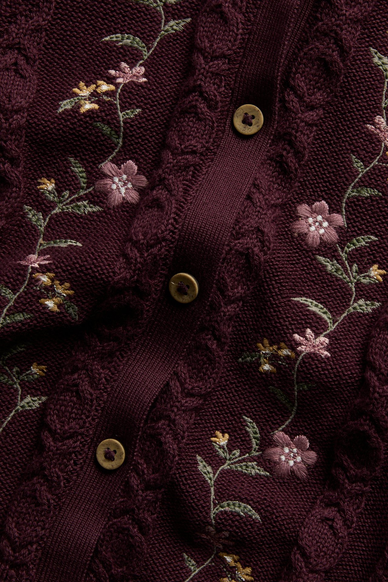 Nahaufnahme einer burgunderfarbenen Zopfstrickjacke mit Blumenstickerei und Holzknöpfen.
