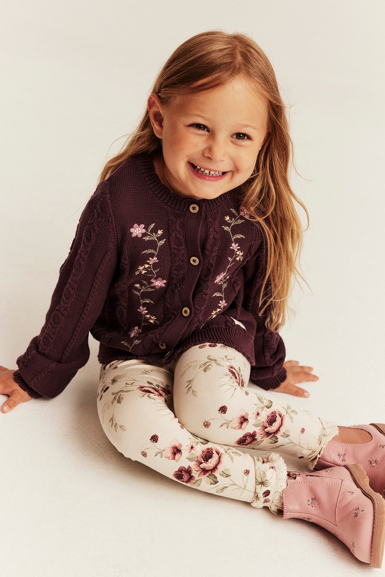 Frontansicht eines Mädchens in dunkelvioletter Zopfstrick-Blumenstrickjacke, floralen Leggings und pinken Stiefeln.