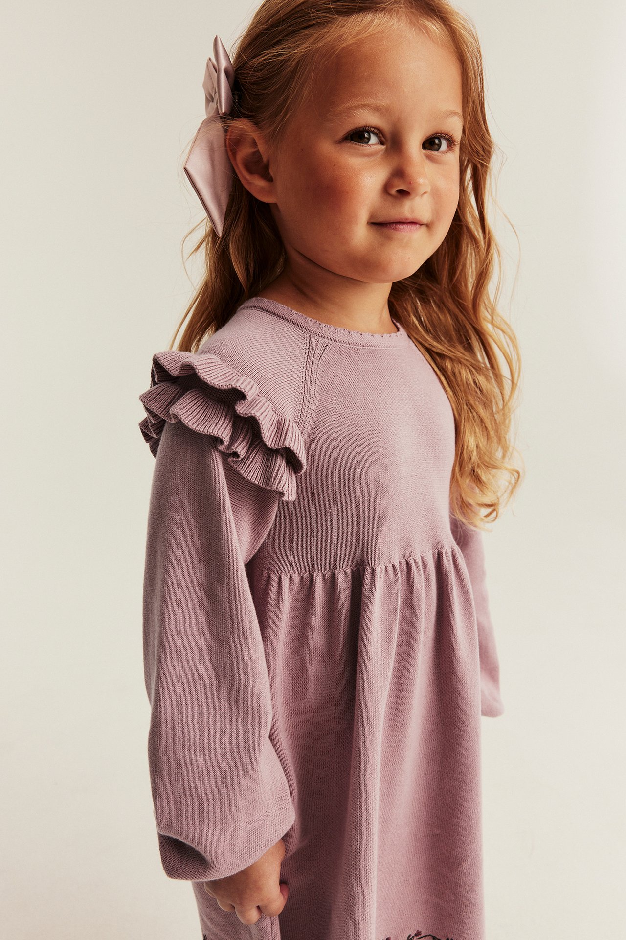 Vorderansicht eines Kindes in lila Strickkleid mit Rüschen und passender Haarschleife.