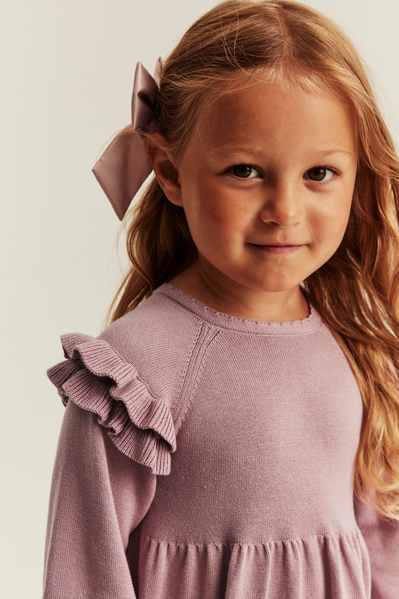 Nahaufnahme Frontansicht eines Kindes in helllila gerüschtem Strickkleid und passender Schleife.