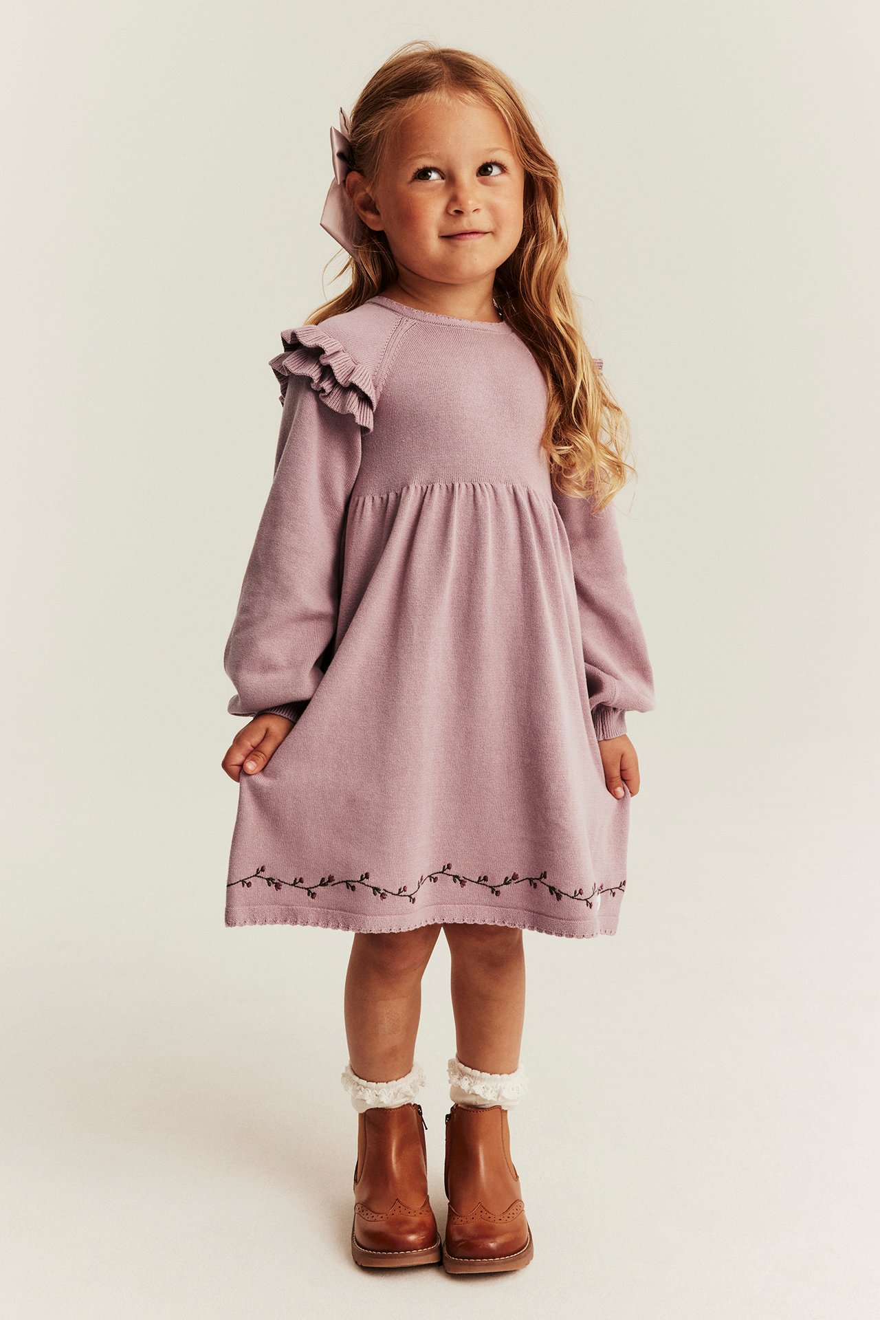 Vorderansicht: Kind in mauvefarbenem Rüschenstrickkleid, besticktem Saum, braunen Stiefeln, weißen Socken, rosa Schleife.