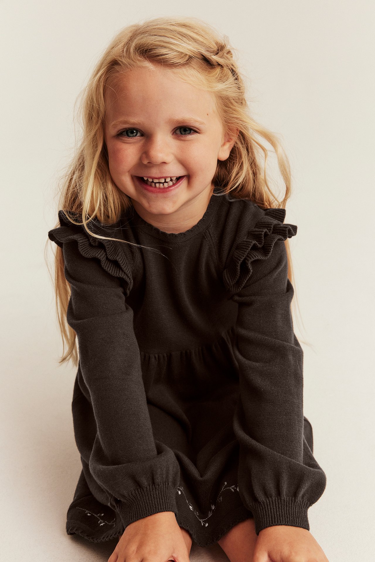 Vorderansicht: Kinderkleid, dunkelgrau, langärmelig, mit Rüschen an den Schultern und besticktem Saum.