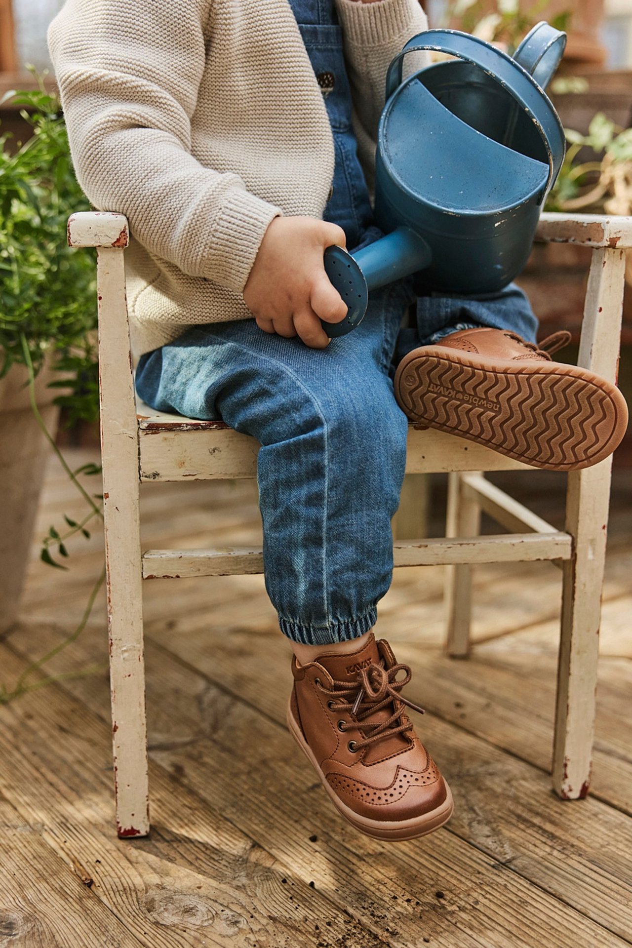 Close-up: Kid in brown leather brogue first walking shoes, blue denim overalls, beige cardigan, holding a watering can.