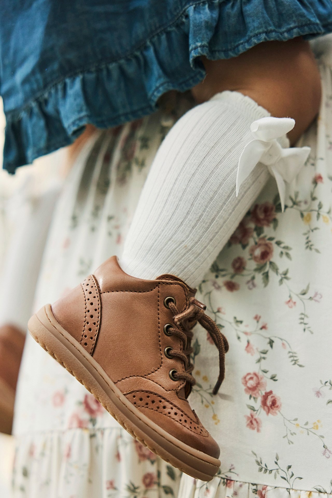 Nahaufnahme eines Babyfußes in braunem Schnür-Lauflernschuh, weißen Rippstrümpfen und Blumenkleid.