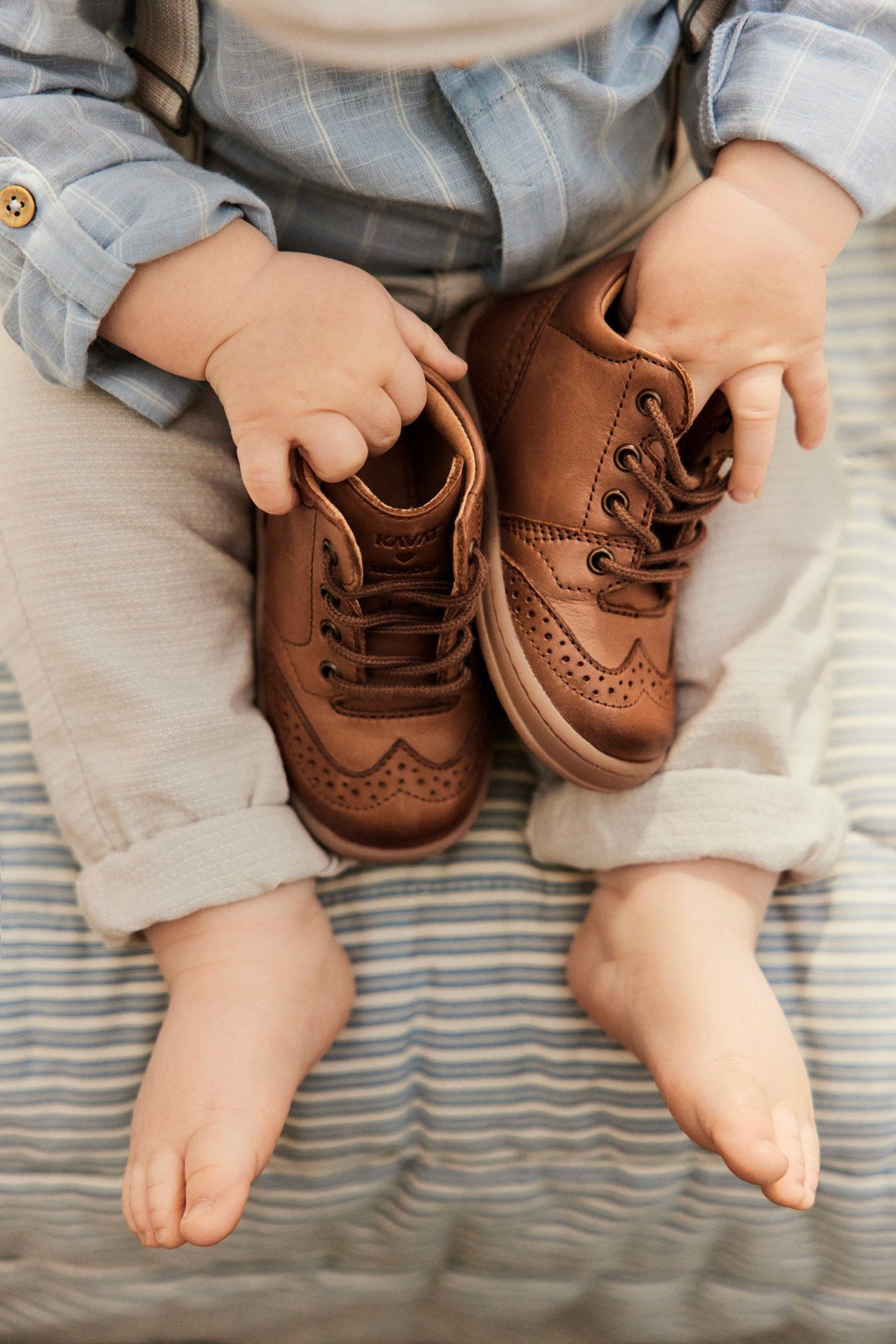 Top-down view of a baby holding brown leather walking shoes with brogue details, wearing a blue striped shirt.