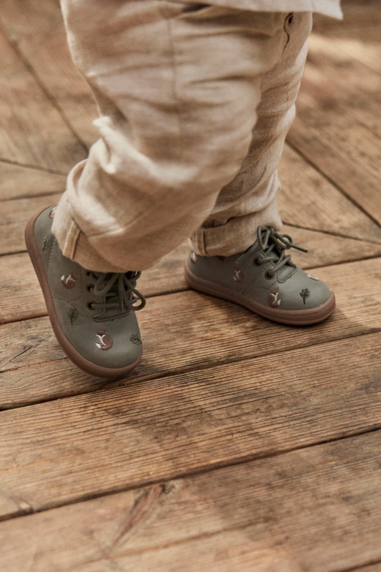 Close-up of a kid's feet in green fox-patterned lace-up walking shoes and beige pants.