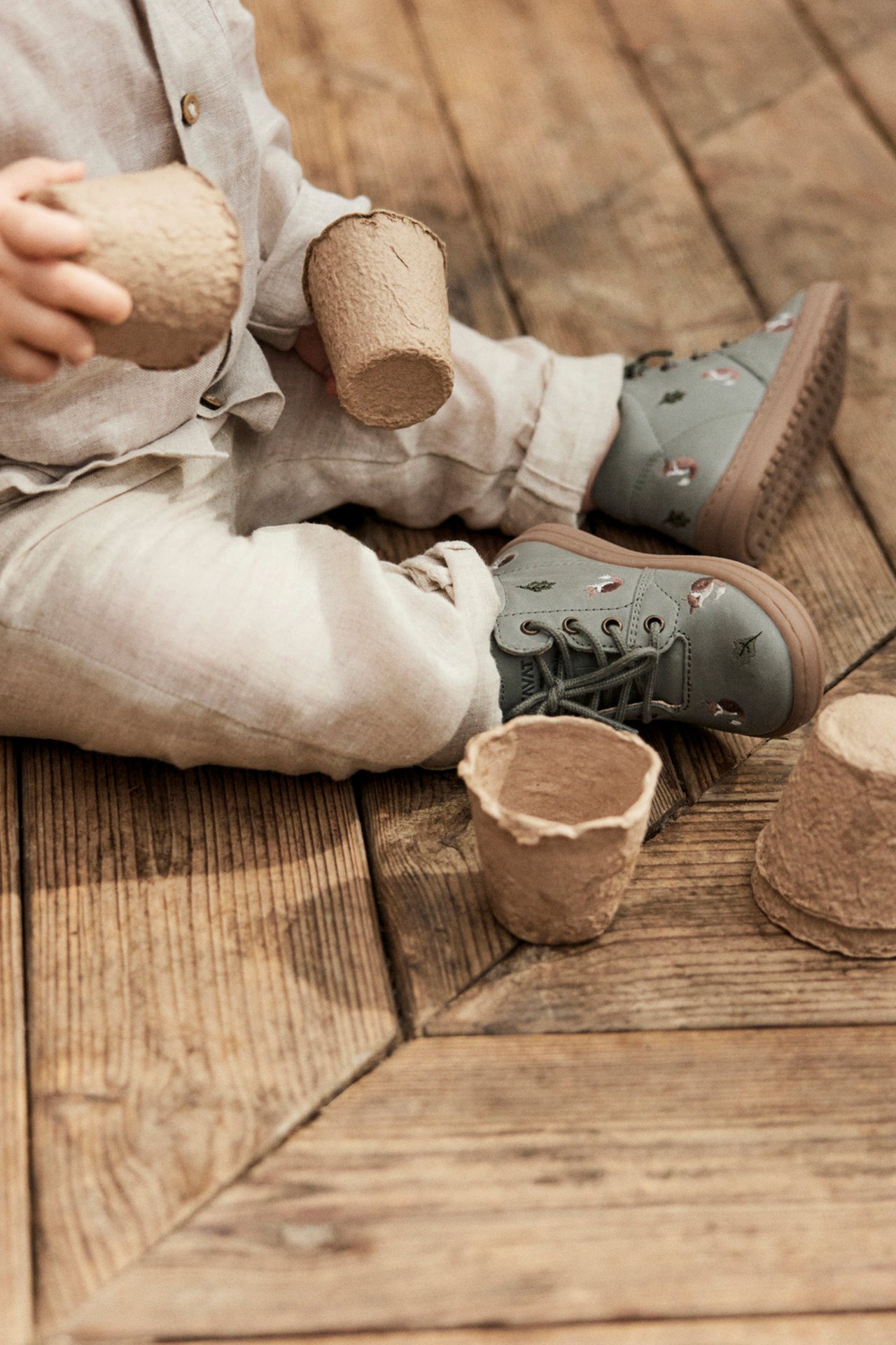 Nahaufnahme eines Babys in beiger Kleidung, das grüne Schuhe mit Tiermuster und Schnürsenkeln trägt und Papiertöpfe hält.