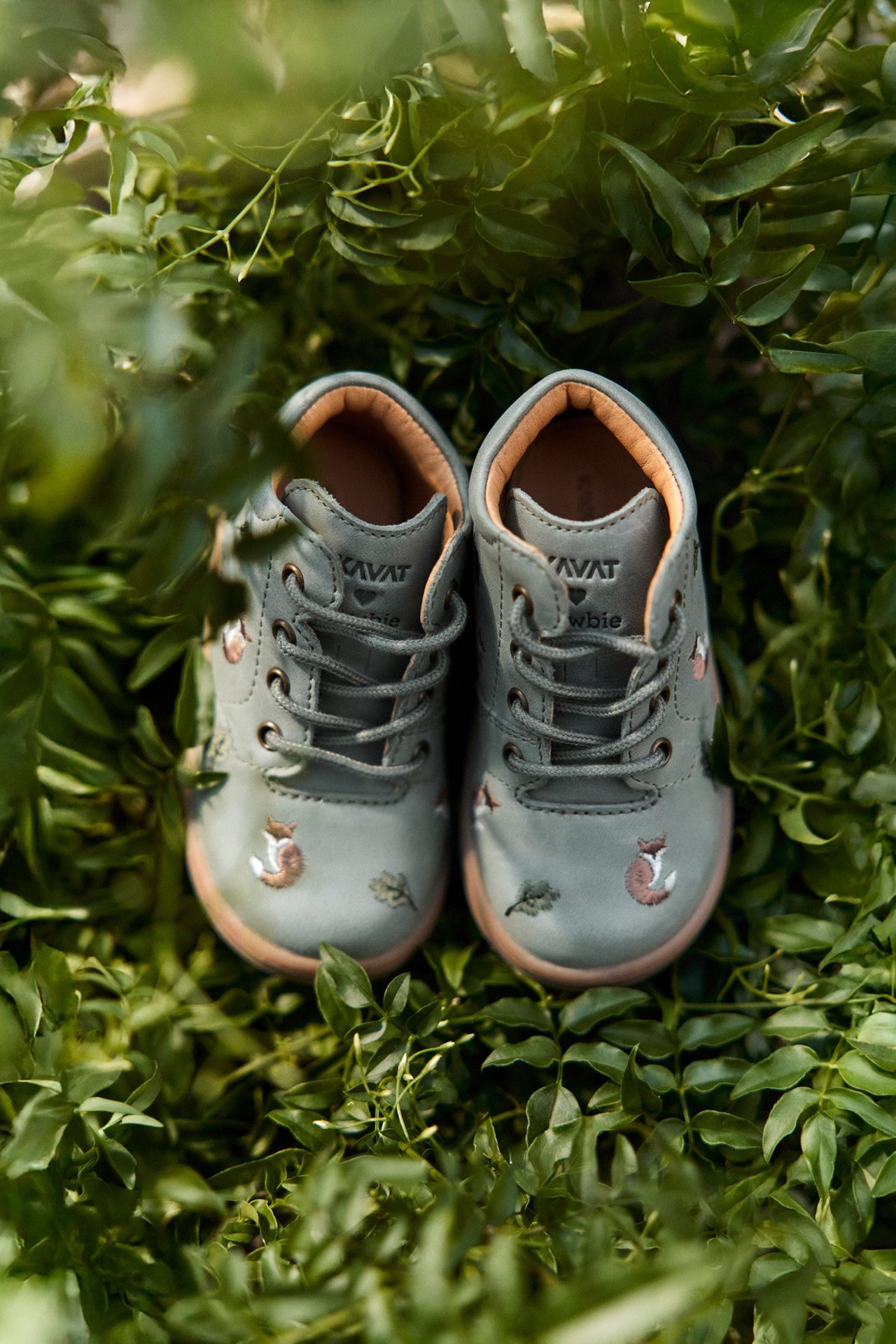 Front view of green leather kid's shoes with fox and leaf embroidery, lace-up closure, nestled in green foliage.