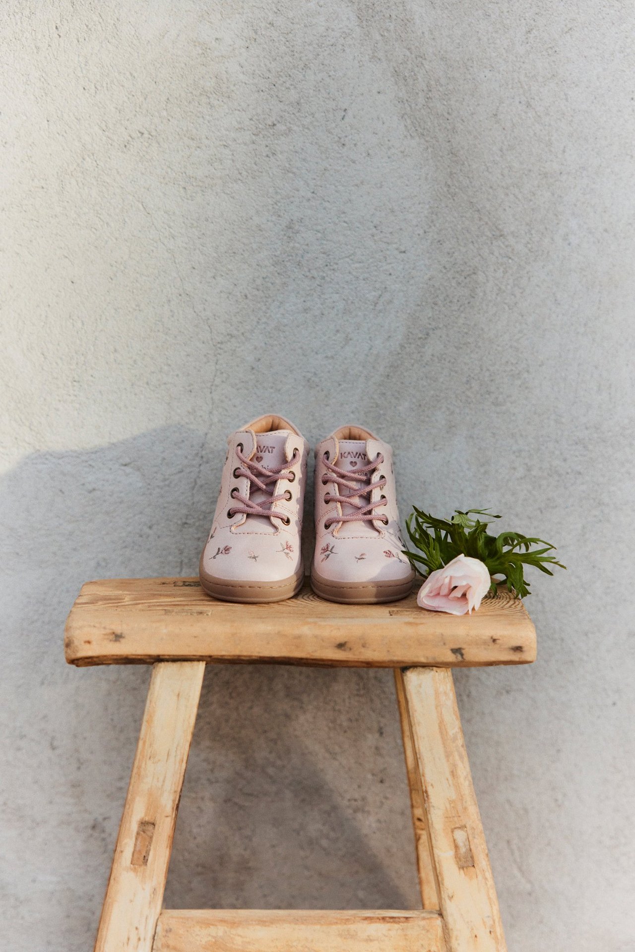 Front-side view of light pink kid's floral patterned lace-up shoes on a wooden stool with a pink flower.