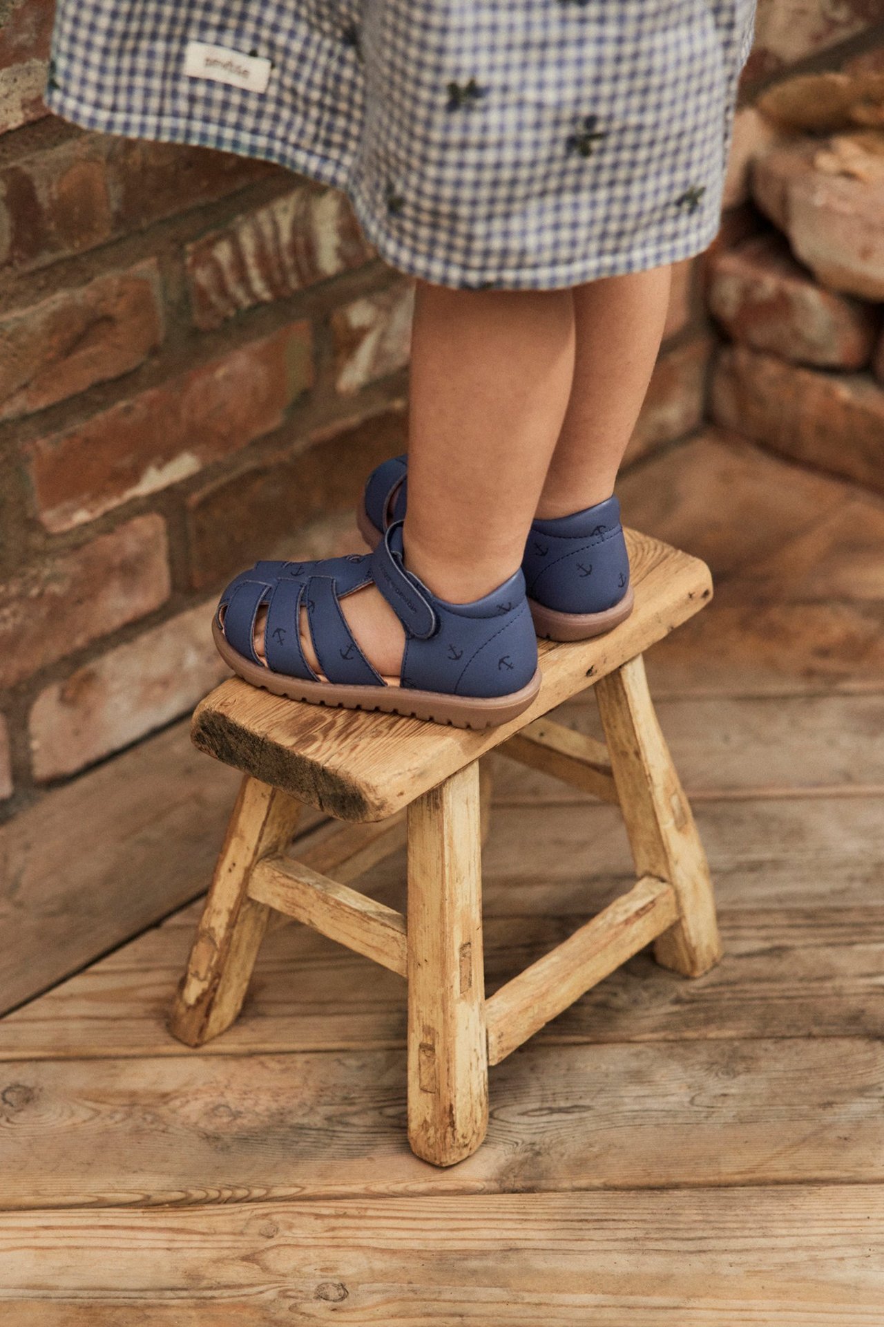 Close-up of a kid wearing navy blue anchor print sandals and a blue-white checked dress.