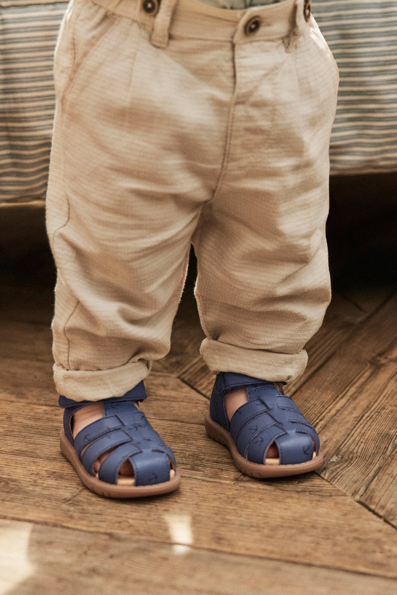 Close-up of a kid wearing blue leather sandals with anchor pattern and textured beige trousers.