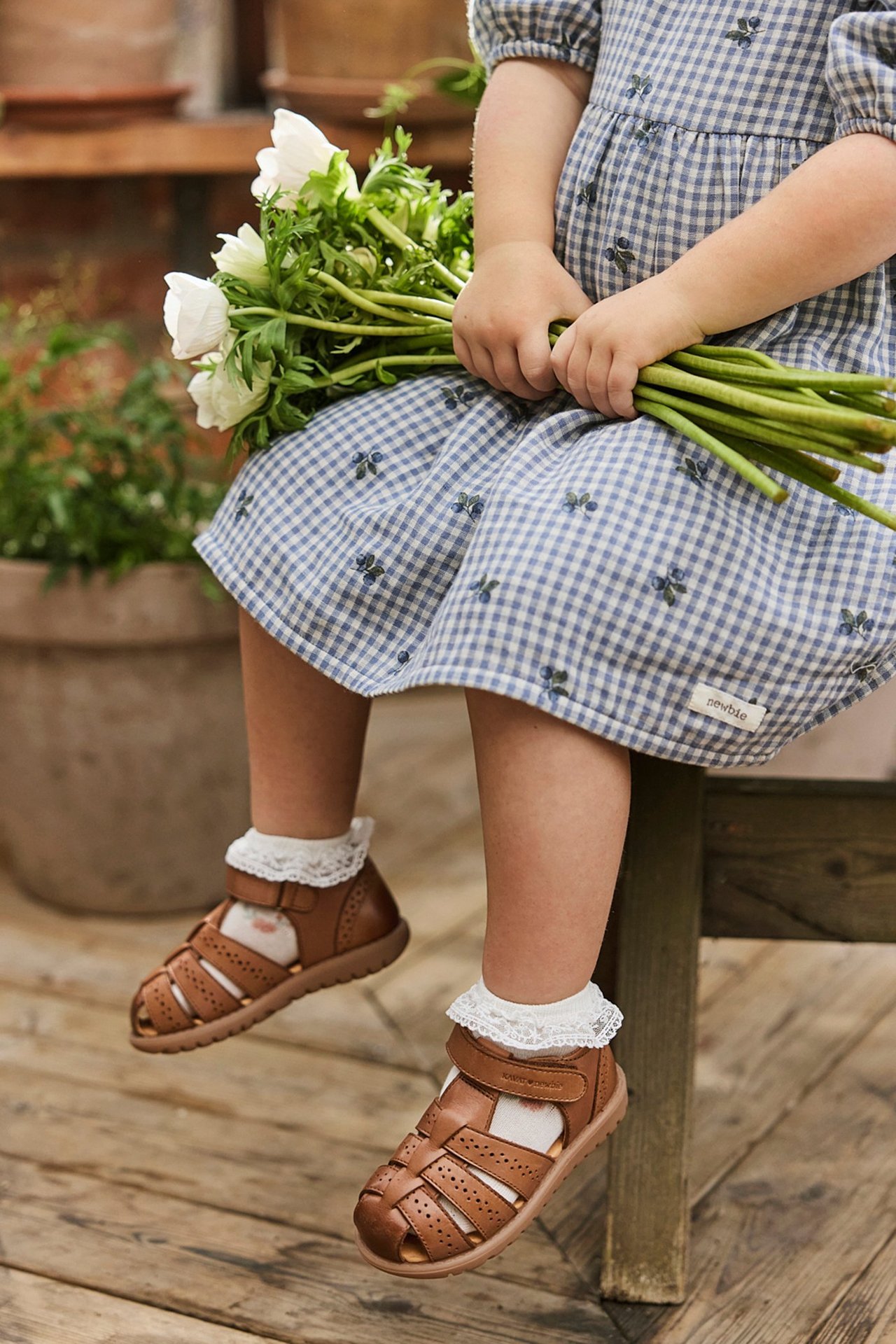 Close-up of a kid wearing brown leather sandals, white lace socks, and a blue gingham dress, holding white flowers.