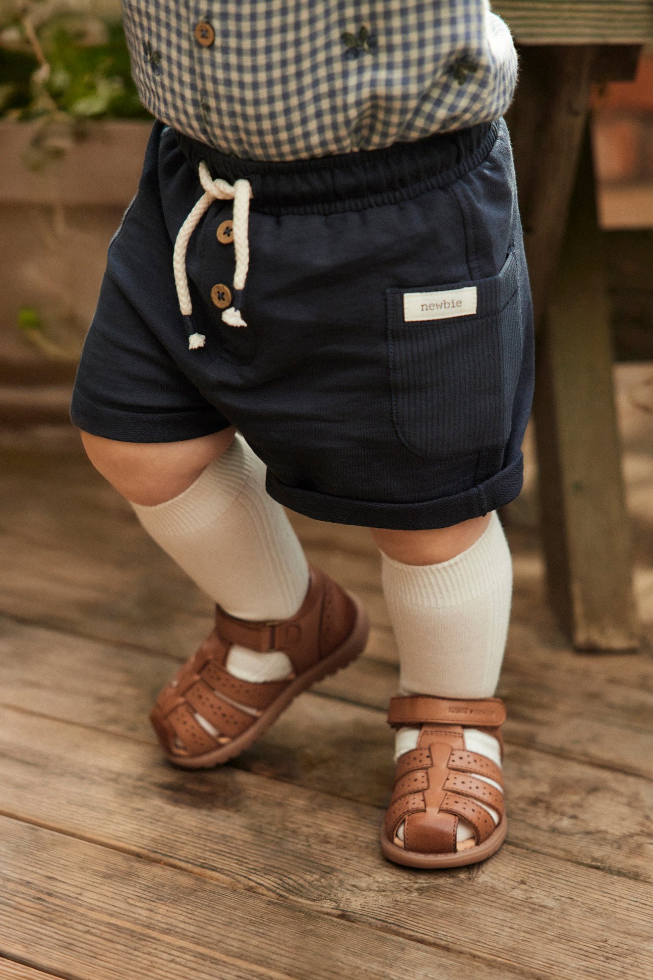 Close-up of baby wearing brown leather sandals, dark blue shorts, a blue checkered shirt, and white socks.