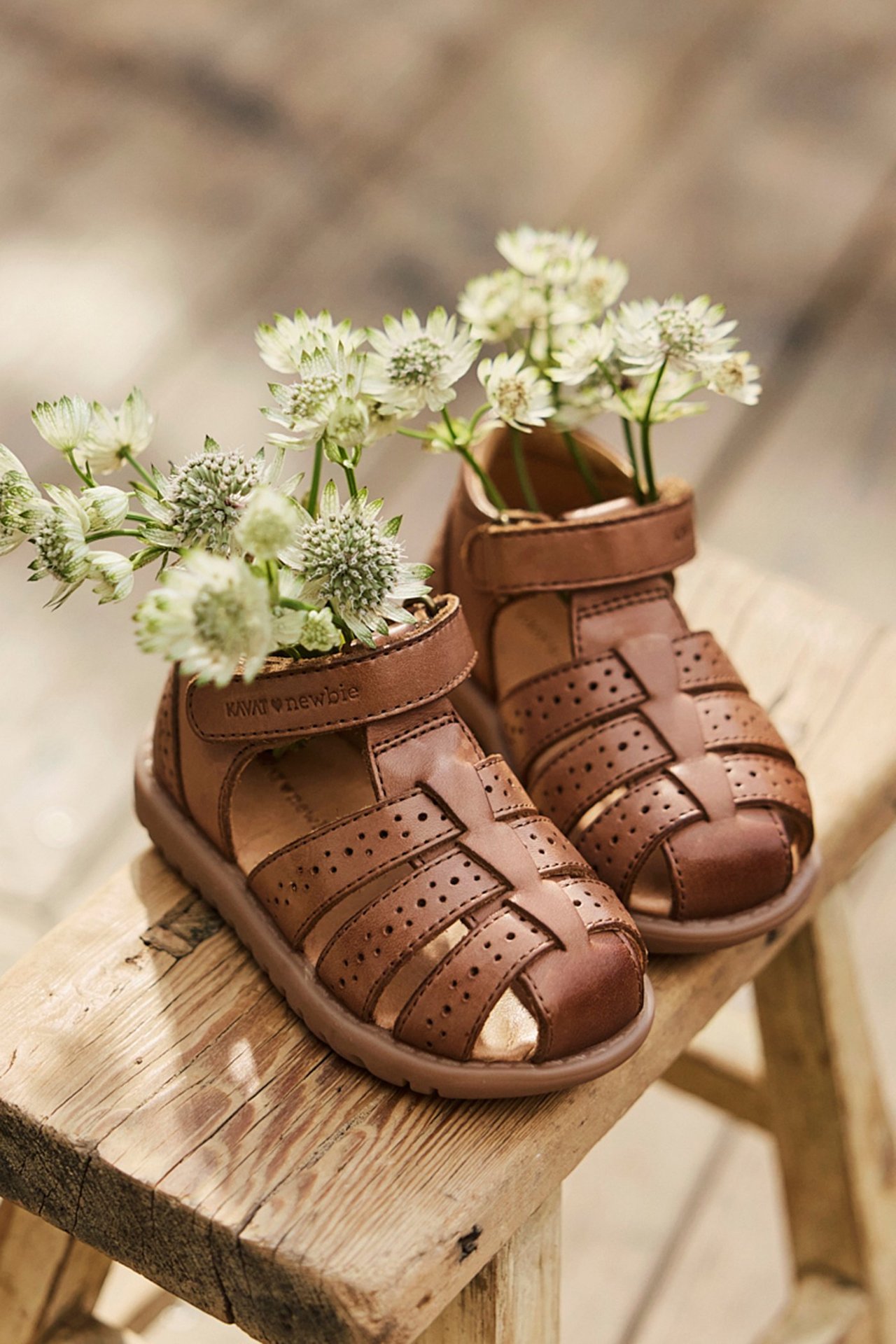Close-up of brown leather kid's sandals with perforated straps, velcro, holding white flowers on wooden stool.