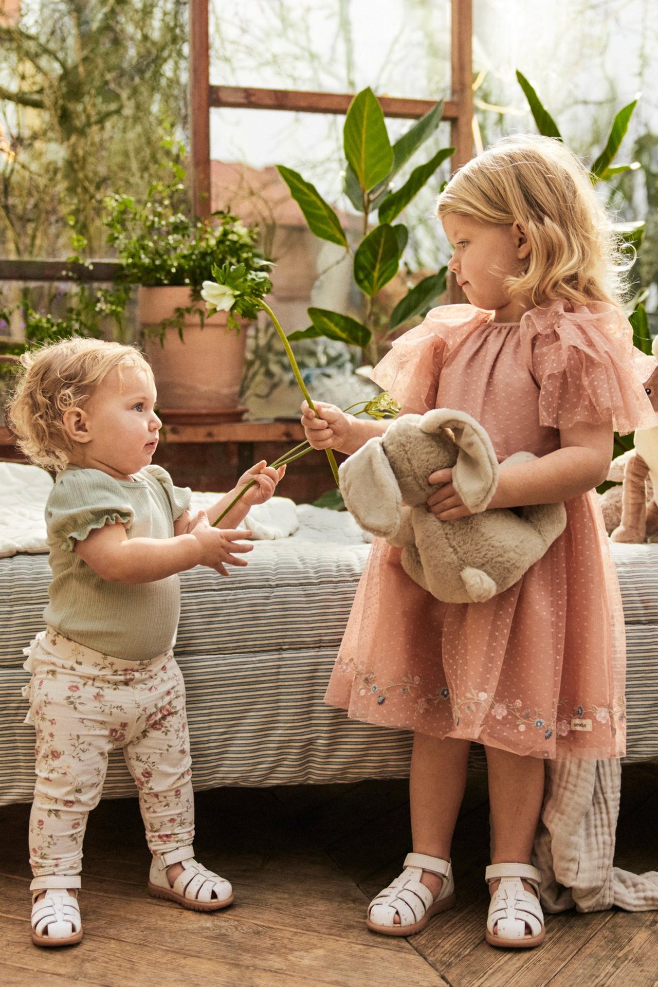 Front view of two kids in white leather sandals. One wears a pink dress, the other a green bodysuit and floral leggings.