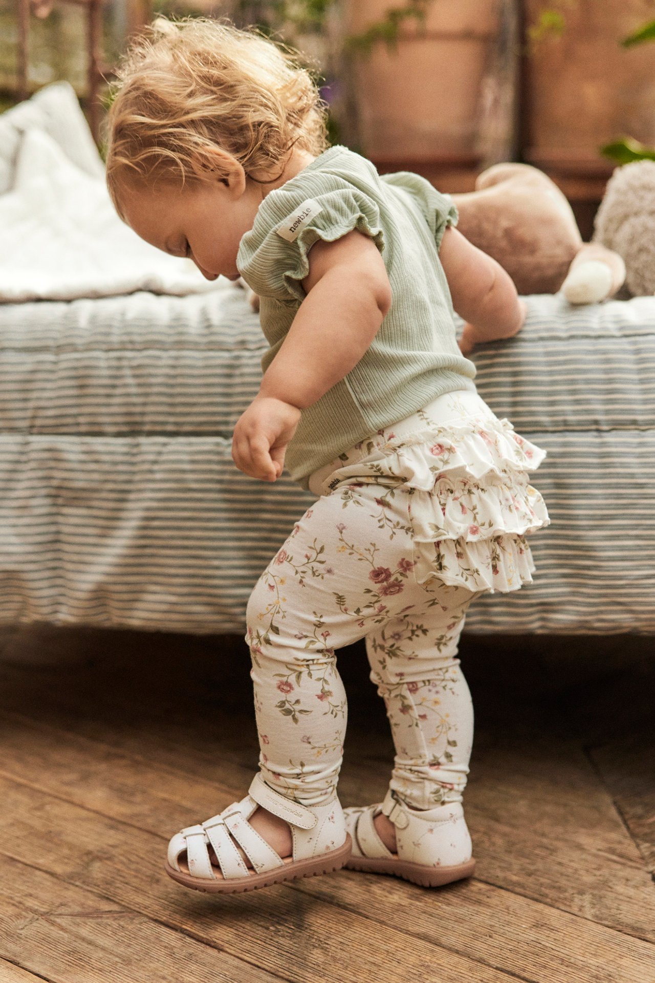 Side view of a baby wearing white floral sandals, a light green top, and cream floral leggings with ruffles.