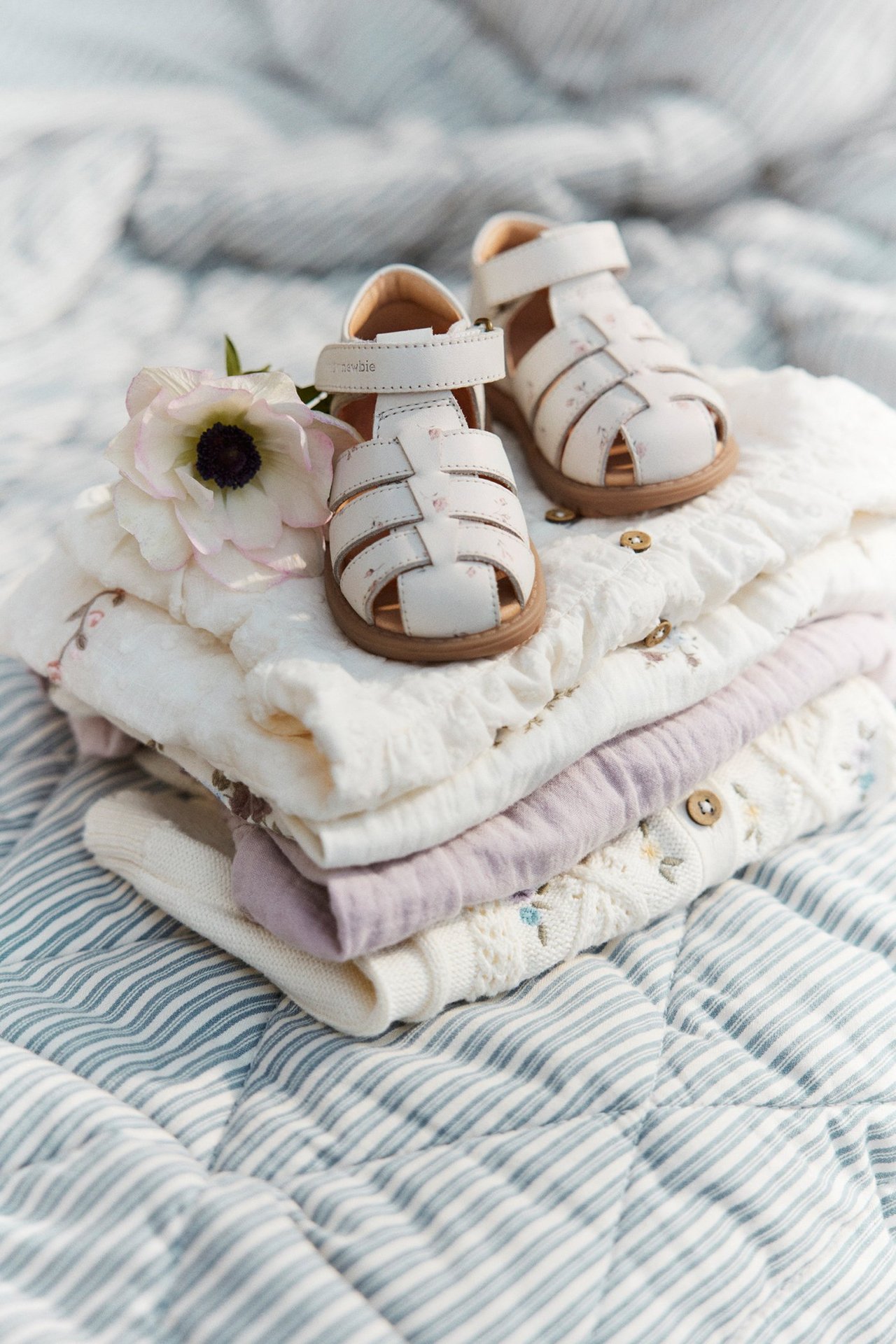 Front view of white floral baby sandals with hook-and-loop fastening, on folded baby clothes and a white flower.