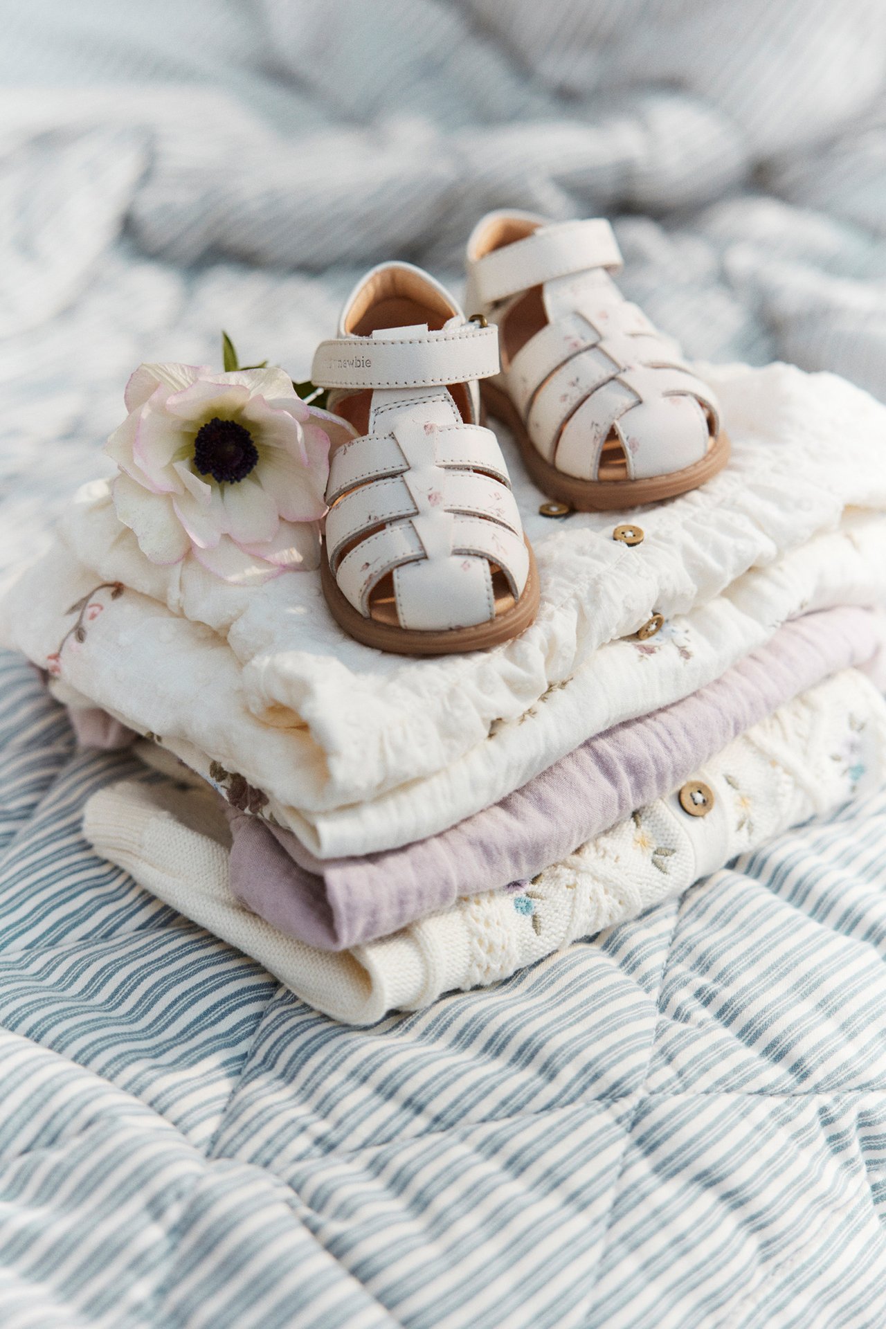 Front view of white floral baby sandals with hook-and-loop fastening, on folded baby clothes and a white flower.