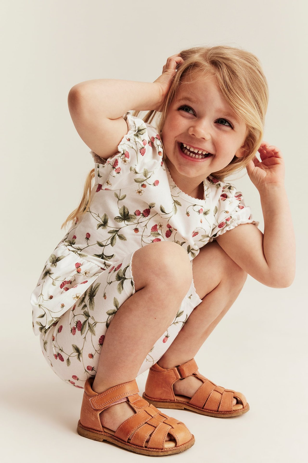 Vorderansicht: Kind in cremefarbenem Erdbeermuster-Peplum-Top und braunen Ledersandalen.