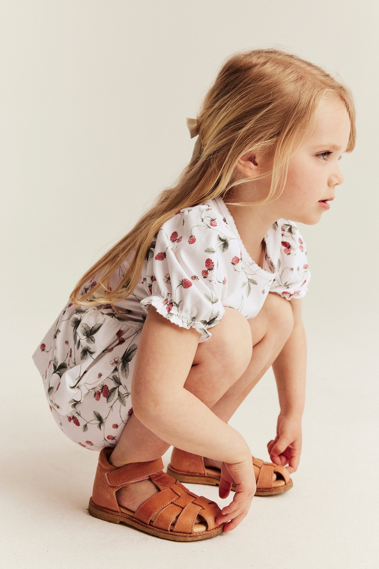 Side view: Kid in offwhite strawberry-patterned peplum top, brown leather sandals, and a hair bow.