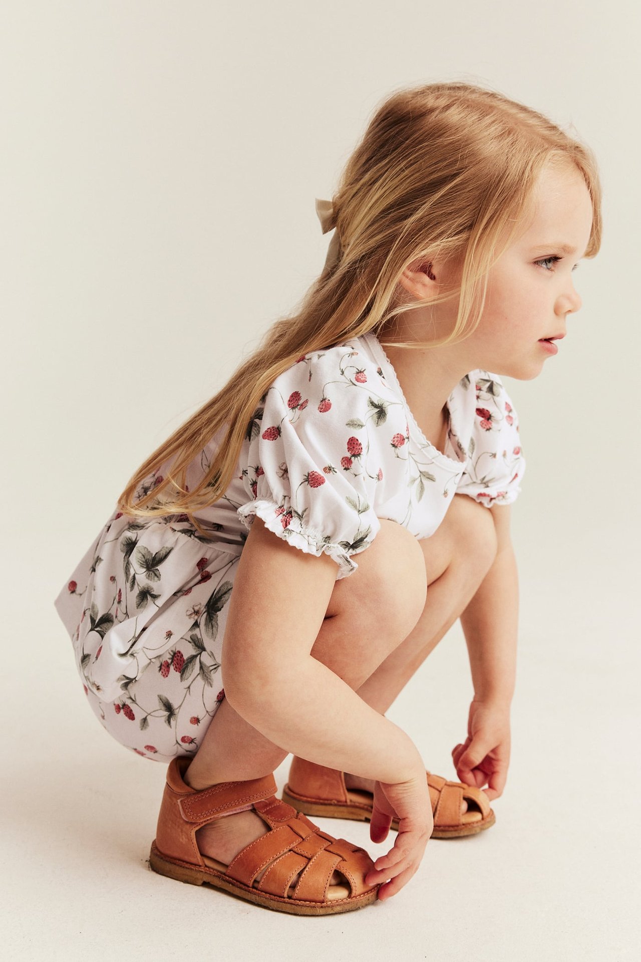 Seitenansicht: Kind in cremeweißem Erdbeermuster-Peplum-Oberteil, braunen Ledersandalen und einer Haarschleife.