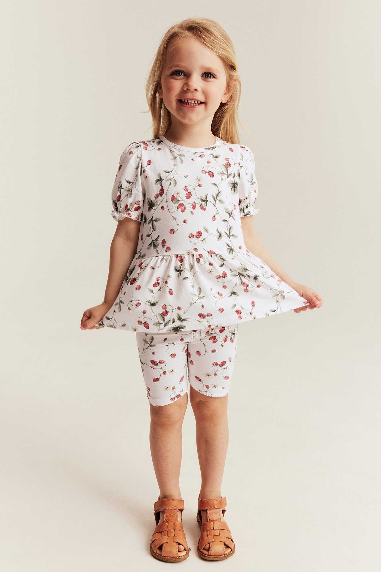 Front view: kid wears an offwhite strawberry-patterned peplum top, matching shorts, and brown sandals.