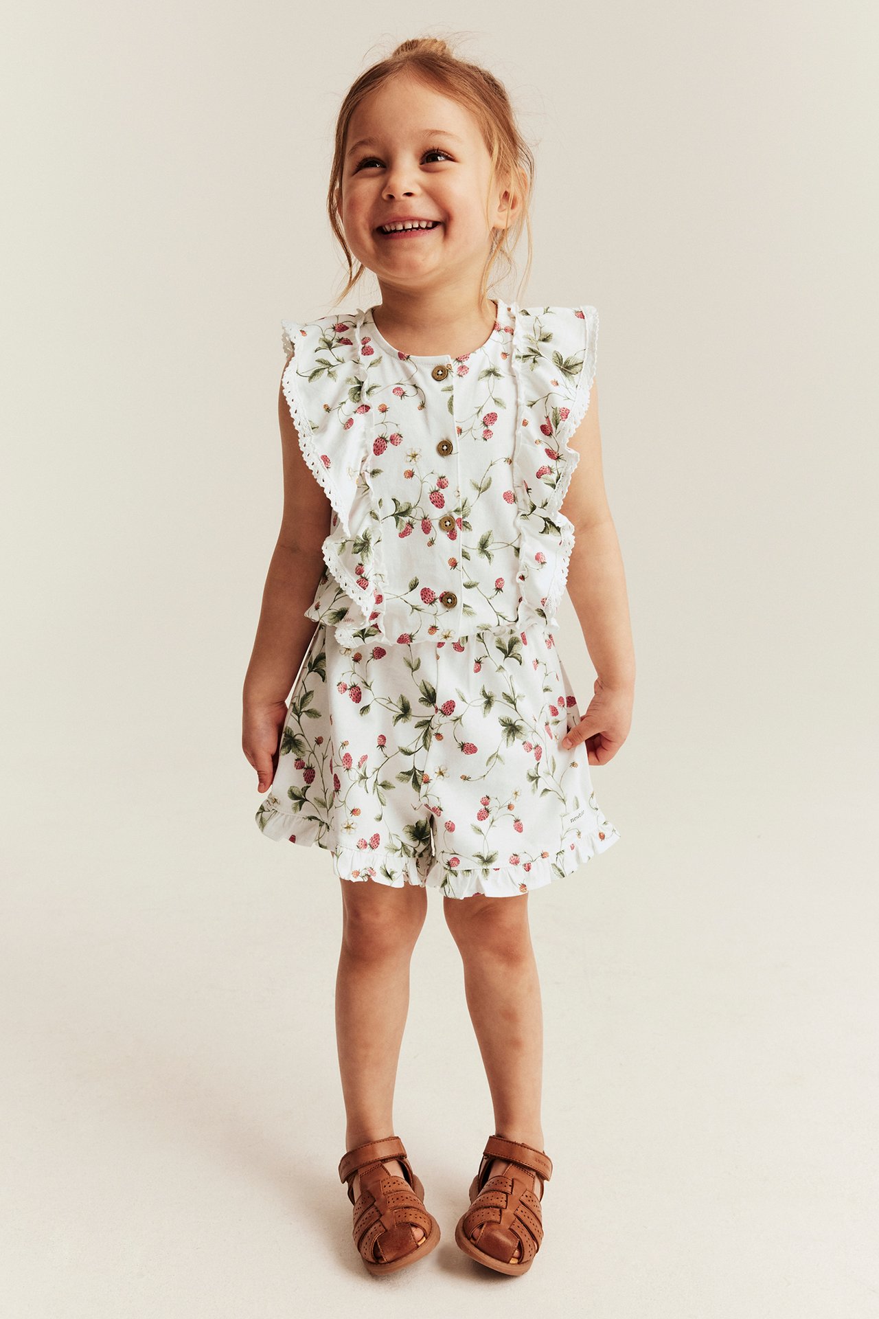 Vorderansicht: Lächelndes Kind in cremeweißem Erdbeermuster-Jumpsuit mit Rüschen und braunen Sandalen.