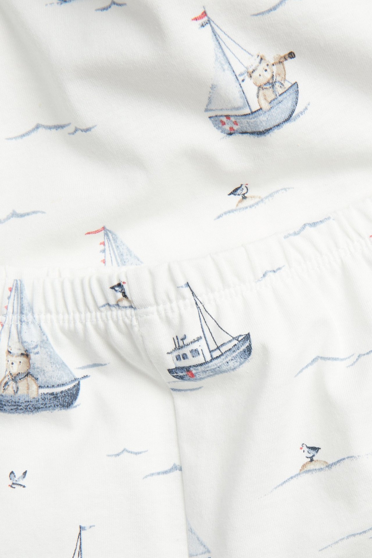 Close-up of off-white cotton pajama fabric with blue sailboats and teddy bear pattern, showing elastic waistband.