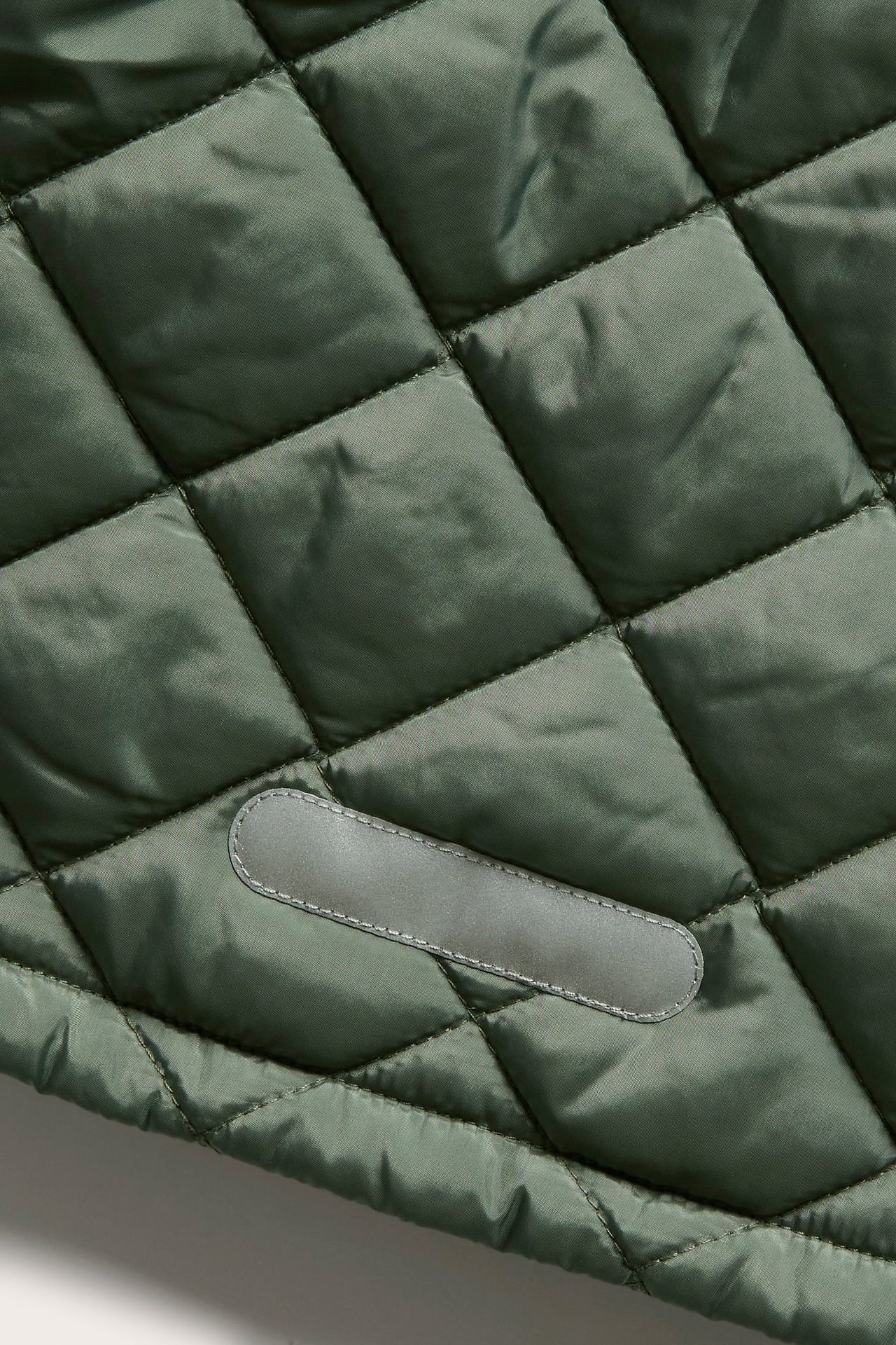 Nahaufnahme einer dunkelgrünen Steppjacke mit Rautenmuster und einem reflektierenden rechteckigen Patch.