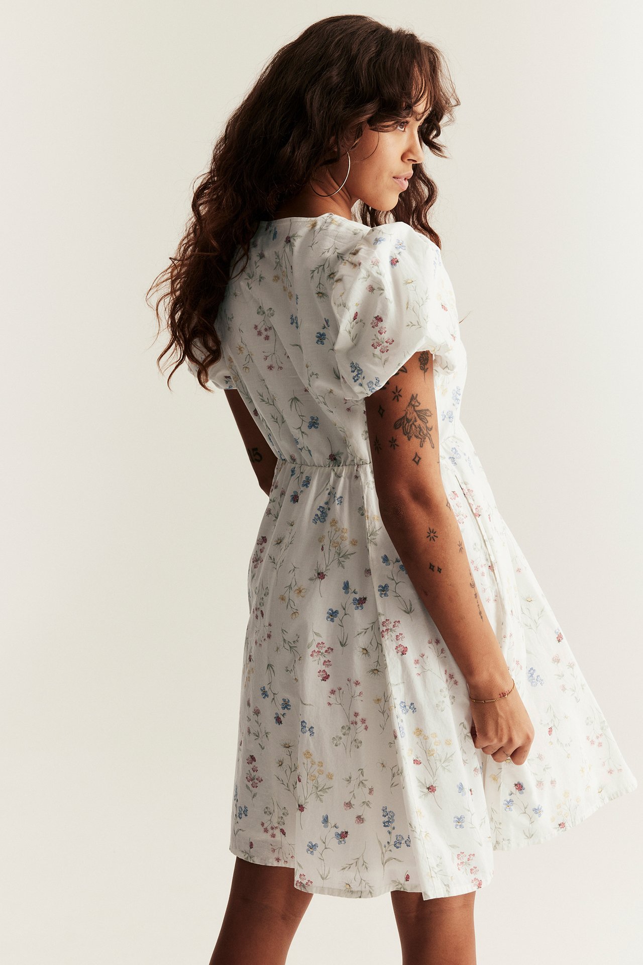 Side-back view of a woman in an off-white floral short dress with puffy sleeves and an elastic waist.