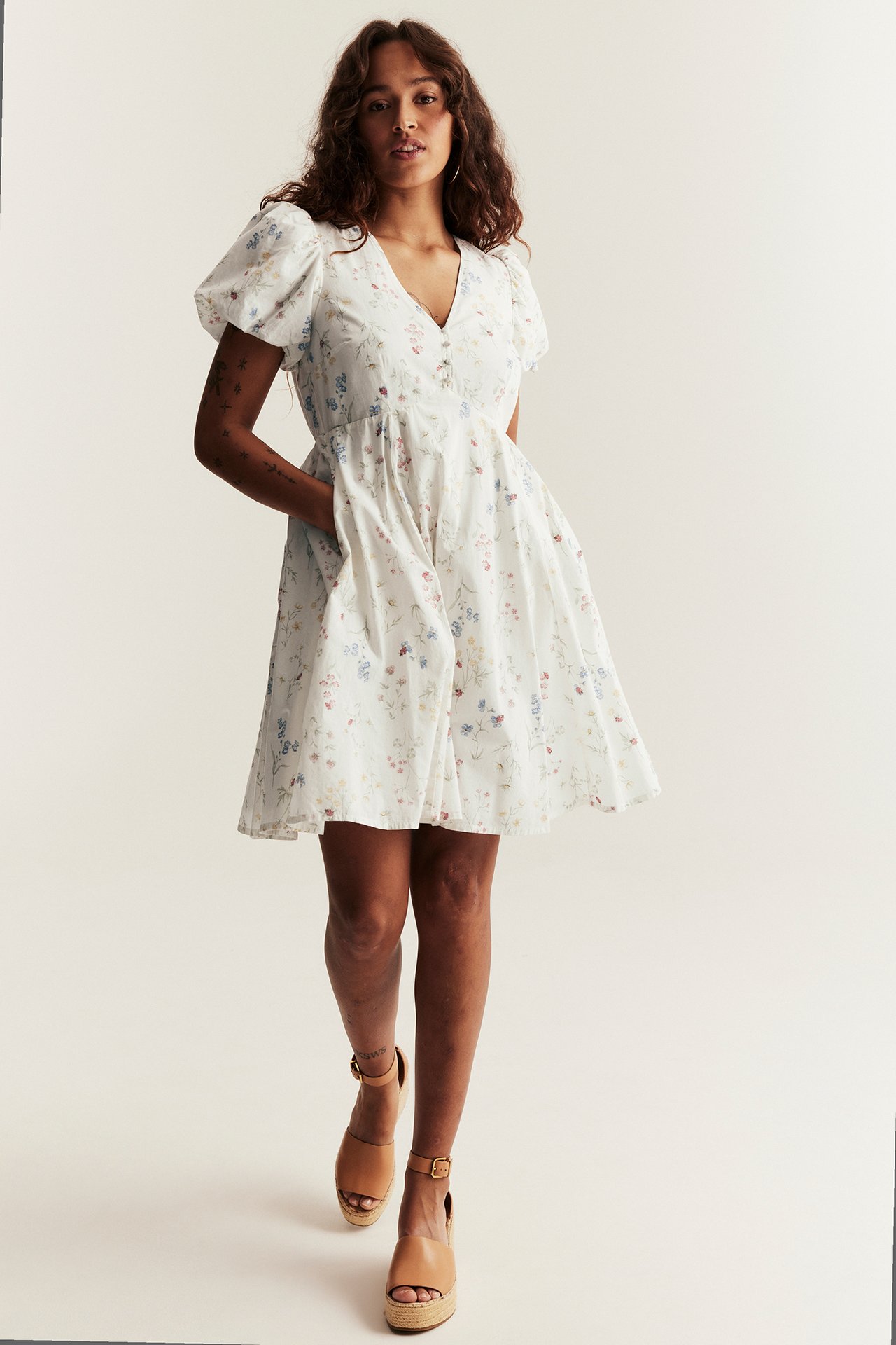Front view of a woman wearing an off-white floral cotton short dress with puff sleeves and tan wedge sandals.