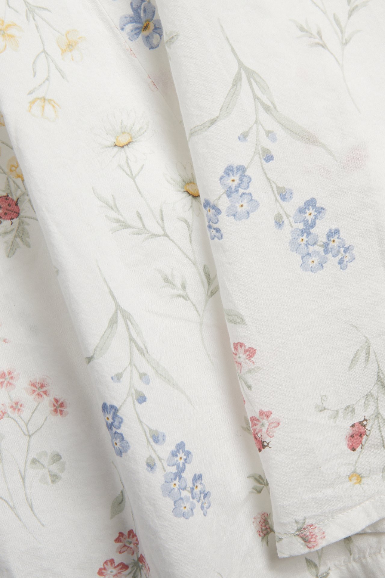 Close-up of off-white cotton fabric with a small floral pattern, featuring blue, yellow, pink flowers, and ladybugs.