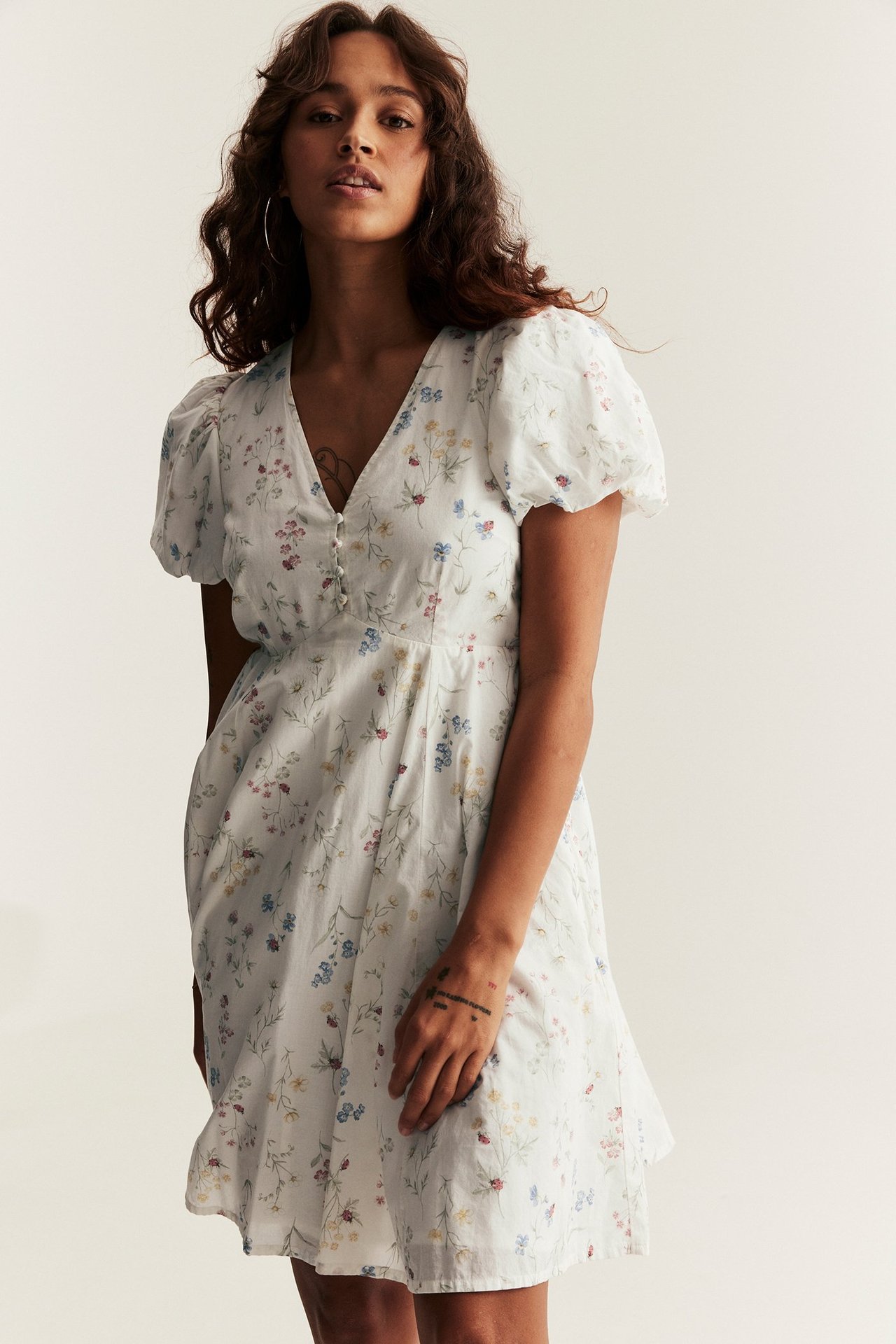 Front view of a woman wearing an off-white floral patterned short cotton dress with puffy balloon sleeves and covered buttons.