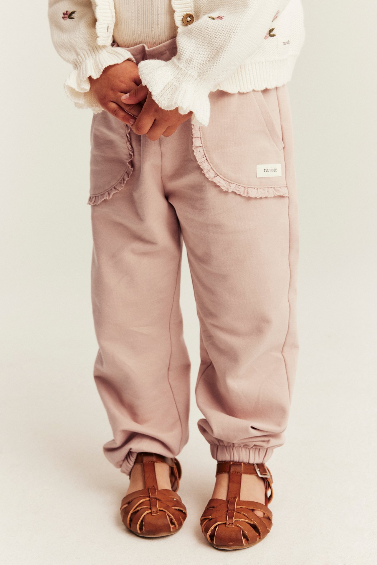 Front view: Kid wears pink cotton jogging pants with lace pockets, a white cardigan, and brown sandals.