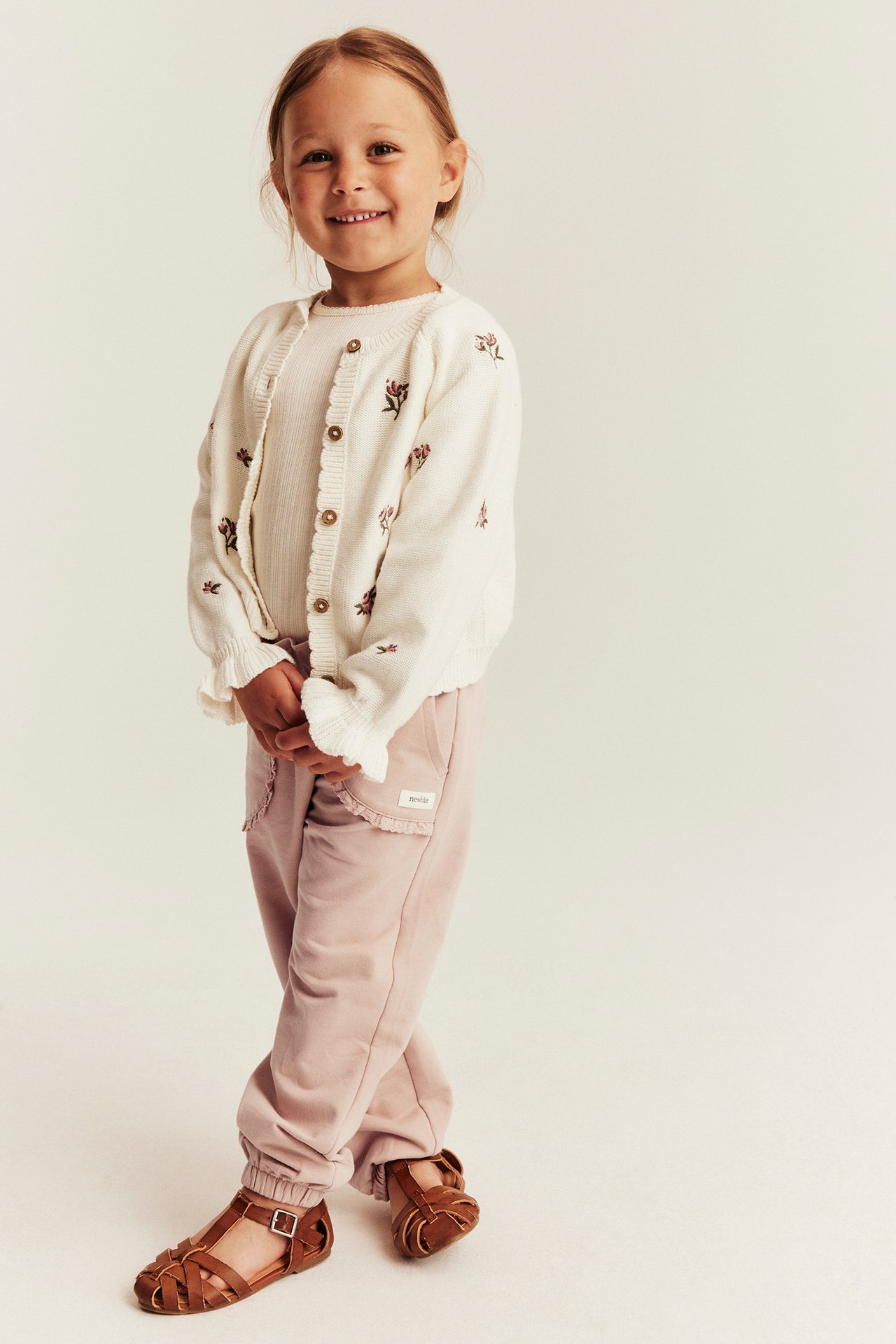 Vorderansicht: Kind in rosa Jogginghose mit Spitze, weißer floraler Strickjacke und braunen Sandalen.