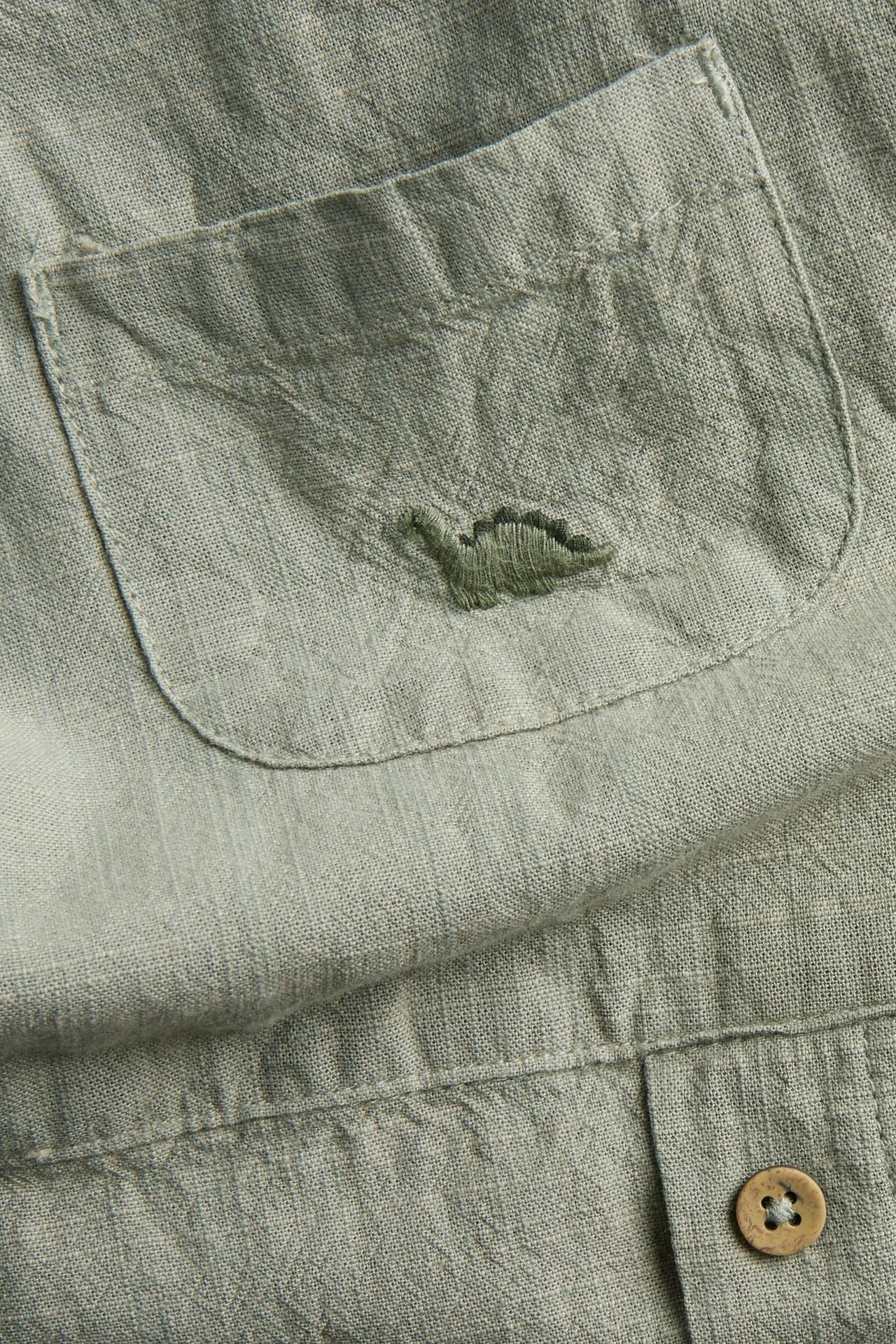 Close-up front of dark green cotton overalls, chest pocket with embroidered dinosaur and a brown button.