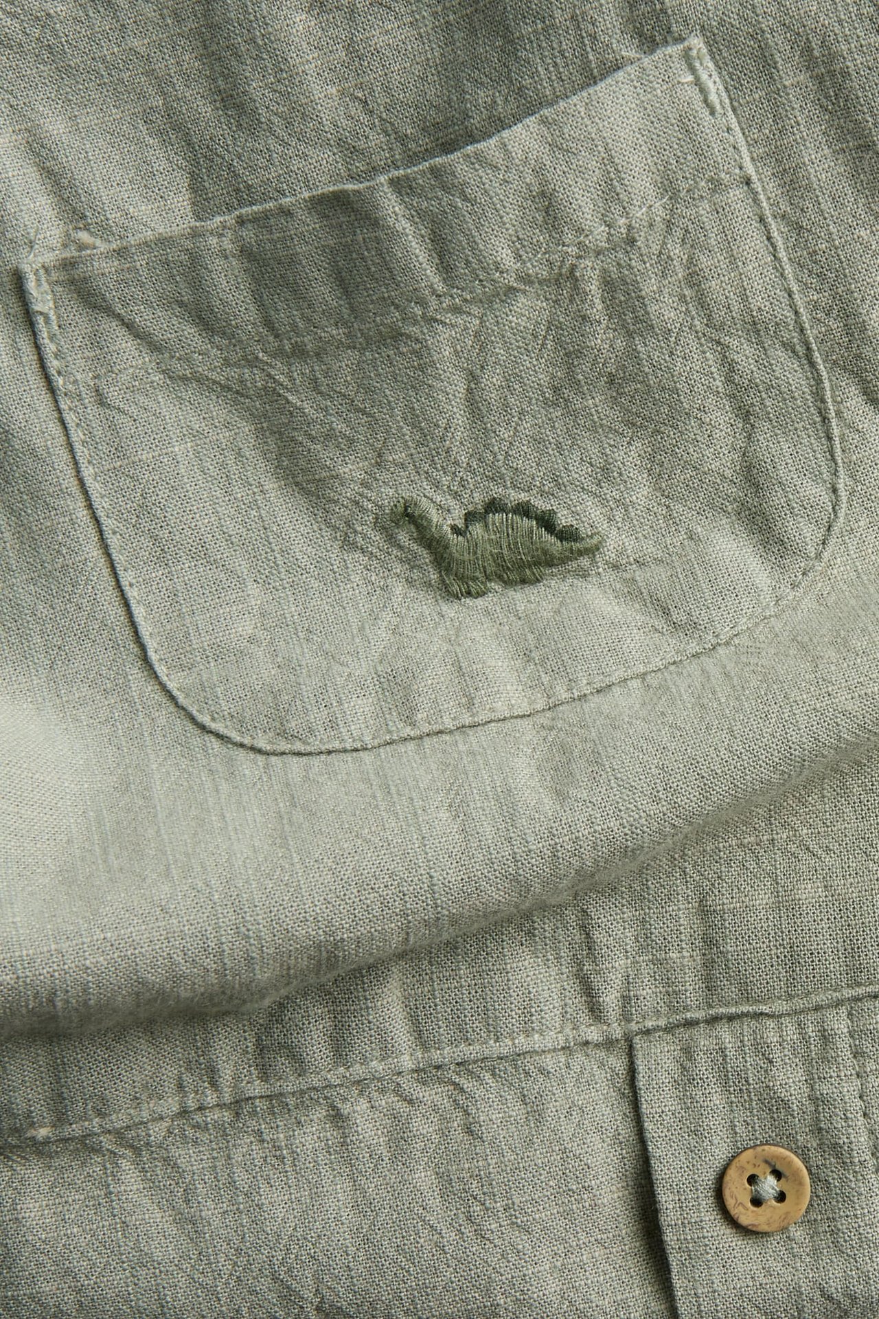 Nahaufnahme vorne von dunkelgrünen Baumwoll-Latzhosen, Brusttasche mit besticktem Dinosaurier und braunem Knopf.