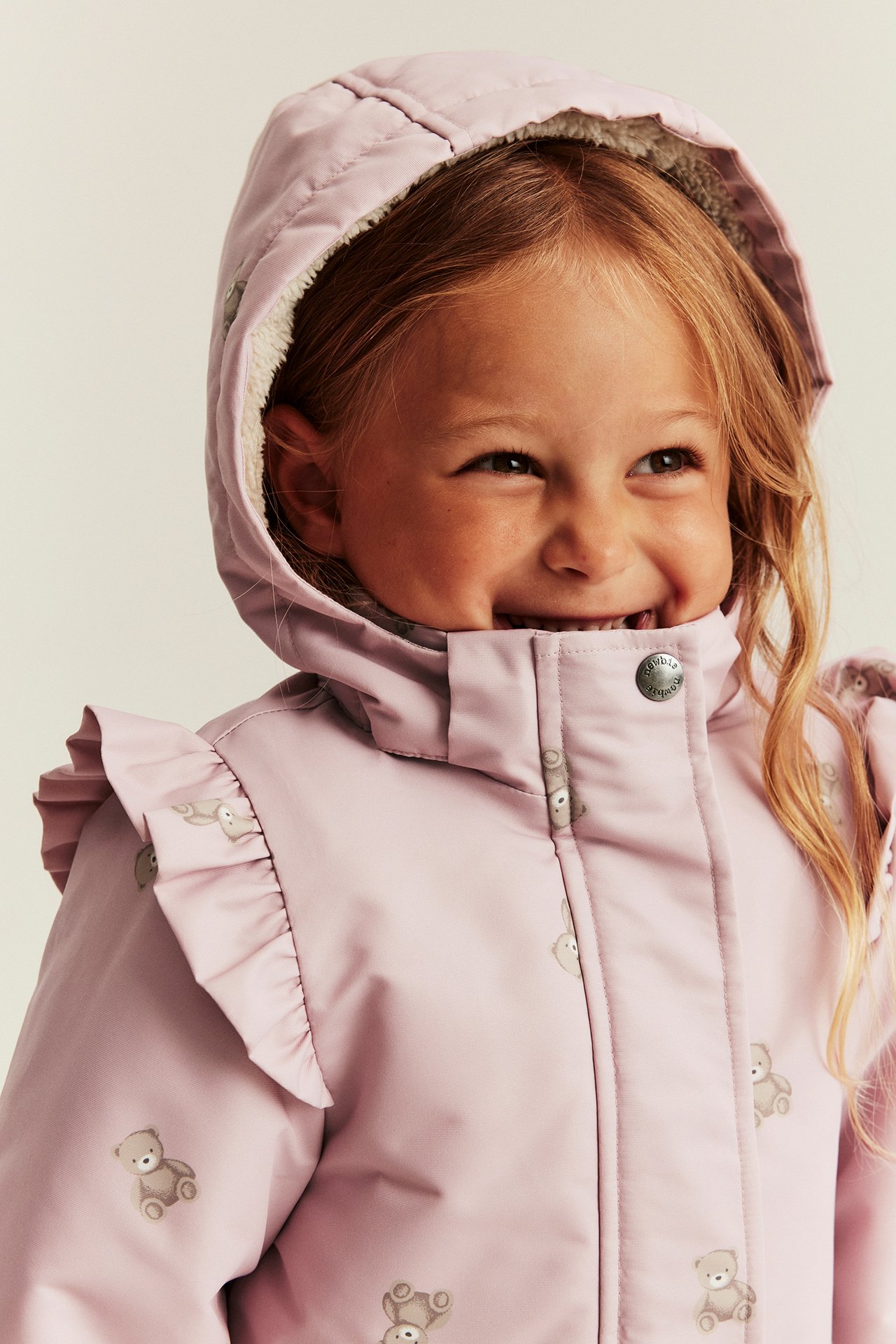 Close-up of a smiling kid wearing a pink teddy bear print hooded jacket with ruffles.