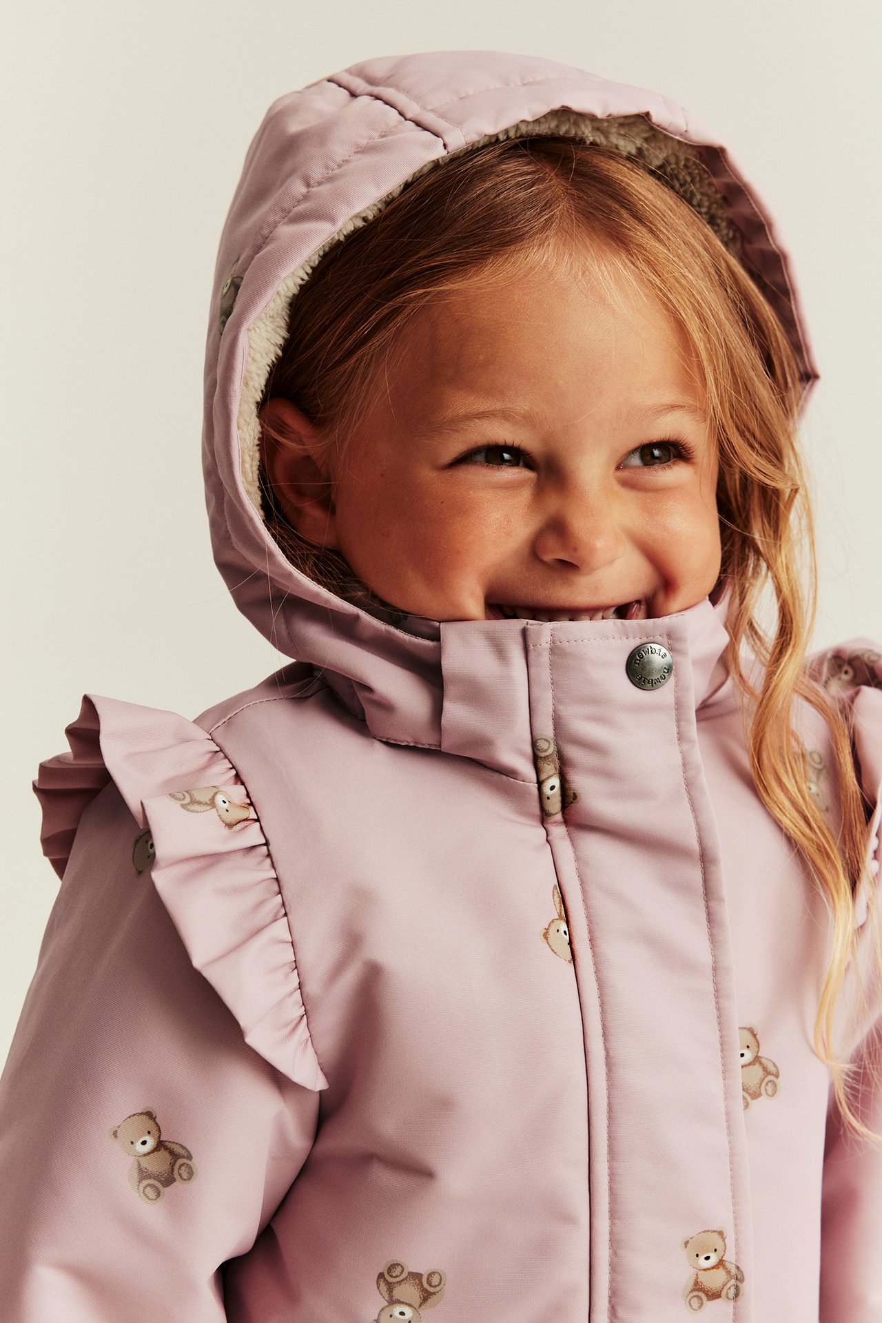 Close-up front view of a smiling kid in a pink teddy bear print hooded jacket with ruffles.